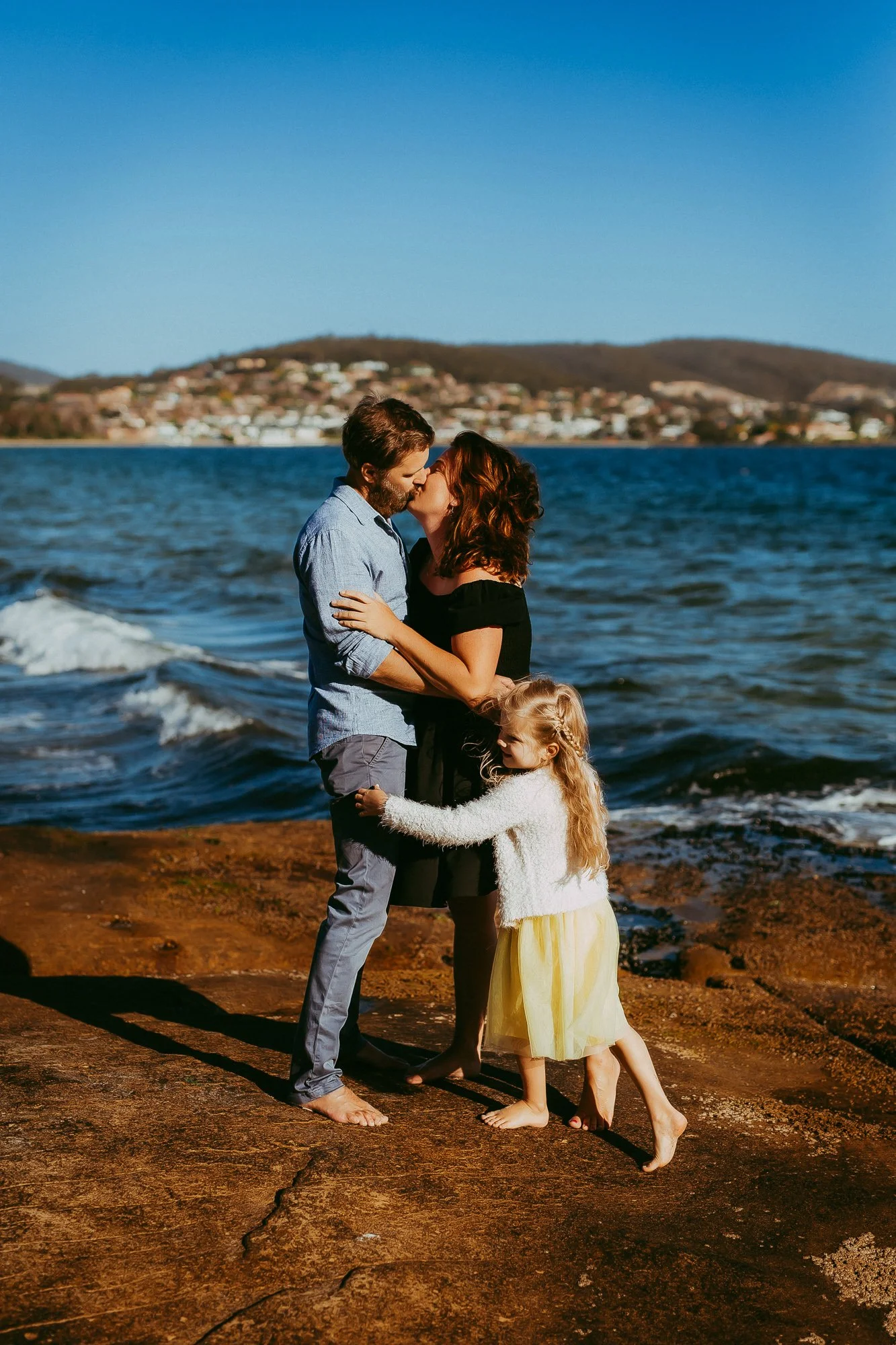 Tasmanian beach family photography session. A couple shares a kiss on the rocks by the water, with a young girl hugging them from the front, at a seaside location under a clear blue sky.