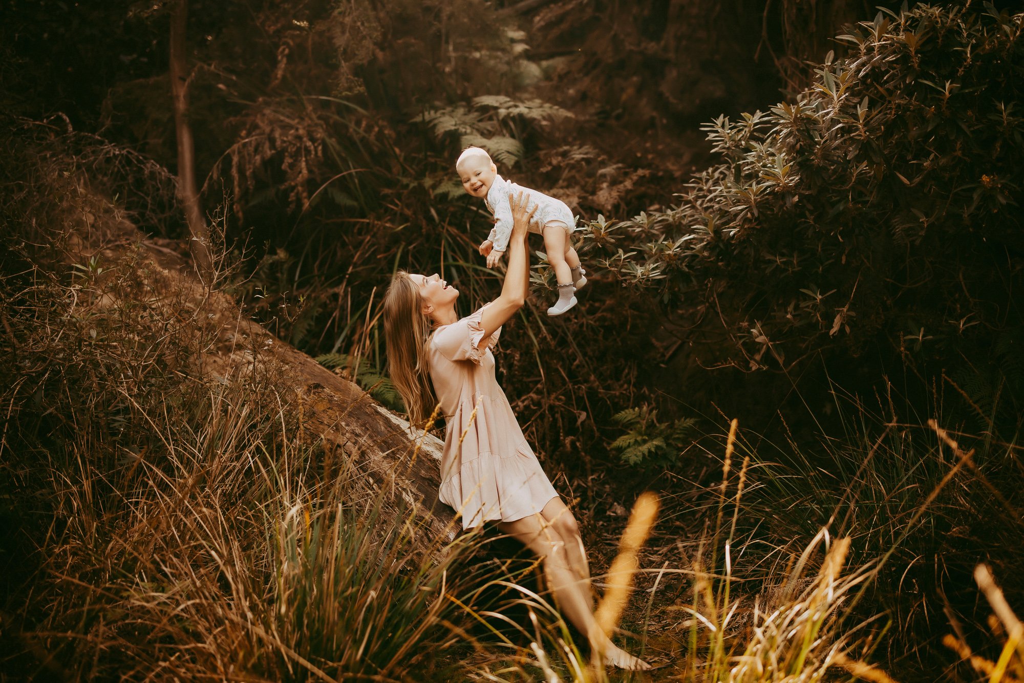 A woman lifting a smiling baby in a forest setting with tall grass and trees around them. Greater Hobart budget friendly family photoshoot.