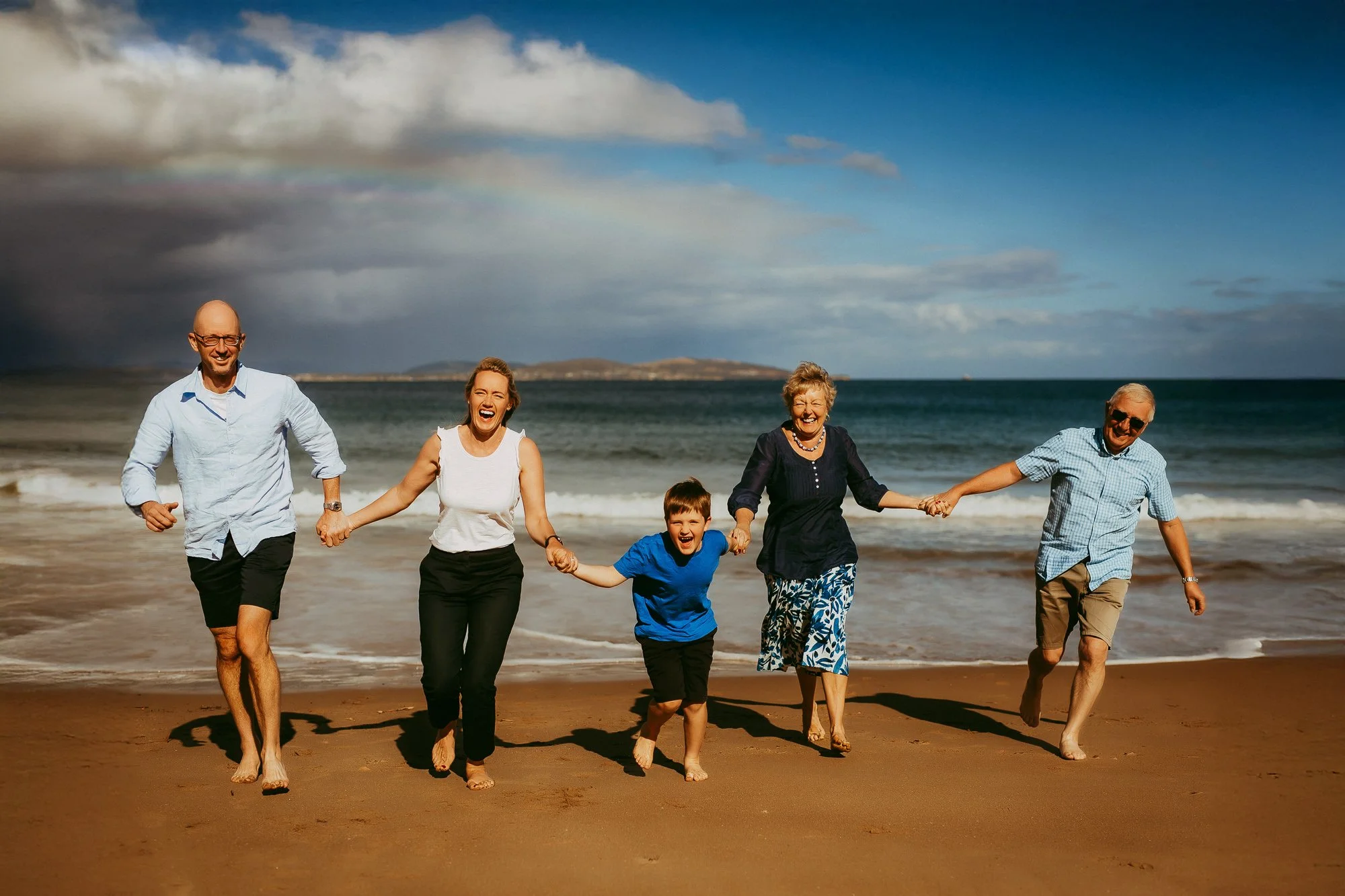 Family of six holding hands and running on the beach with sea and cloudy sky in the background.