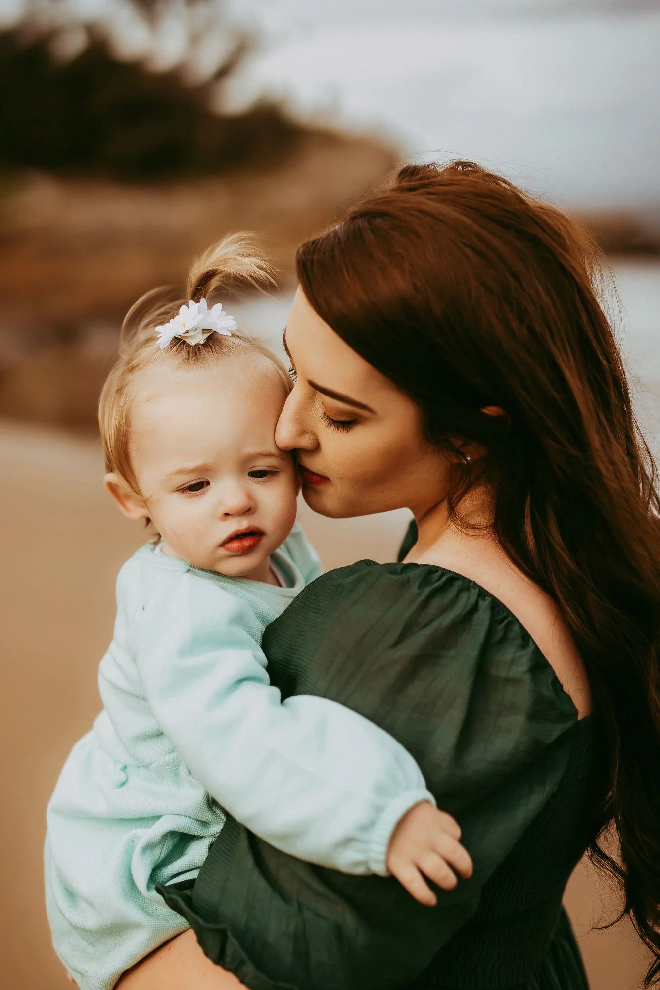 A woman with long brown hair holding a young girl with blonde hair and a white hair bow on a beach. Stunning Tasmanian family photography session.