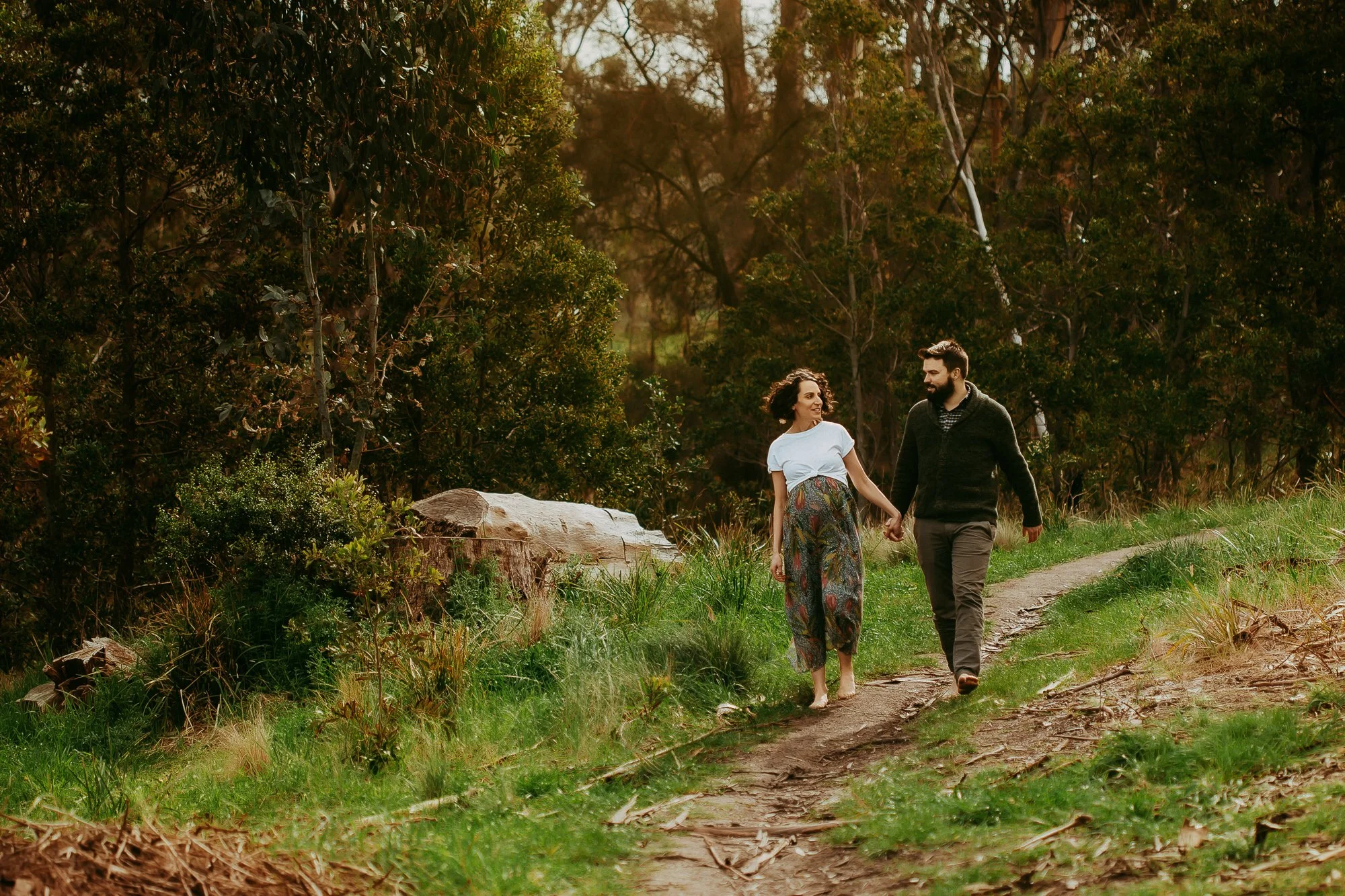 A couple walking hand in hand on a dirt trail through a forested area during daytime.