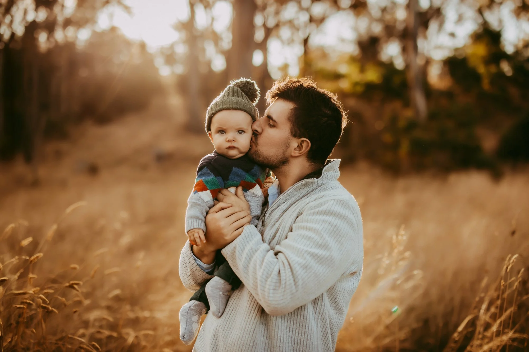 A man holding a young child outdoors during golden hour in a grassy field with trees in the background, with the man kissing the child's cheek. Hobart area golden hour family photoshoot.
