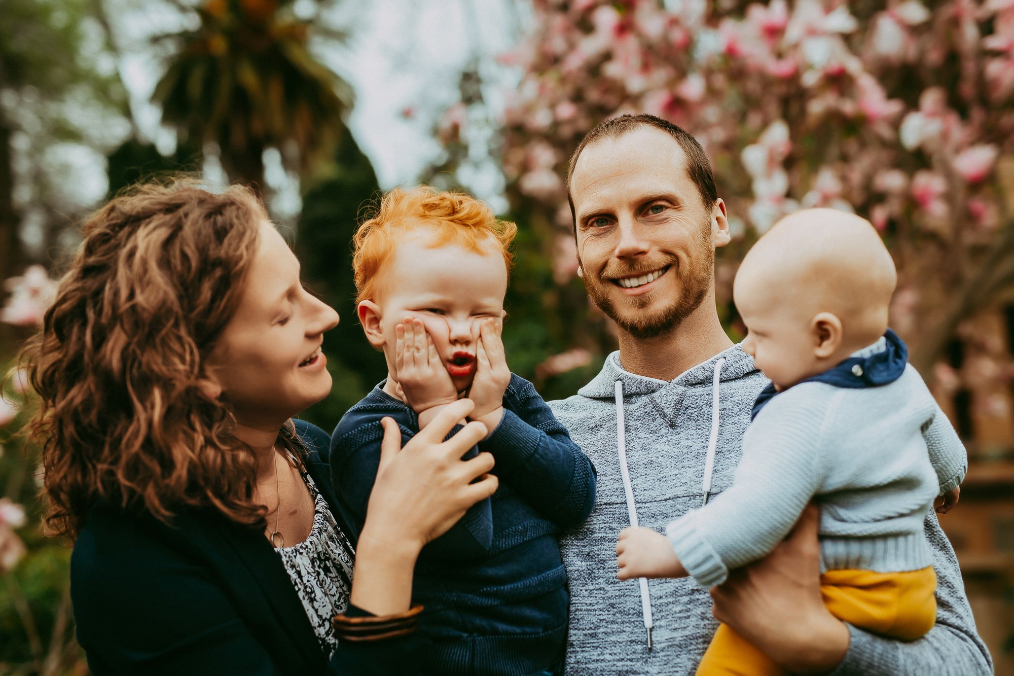 A smiling family of four outdoors with pink blossoms in the background. Unposed Hobart family photography