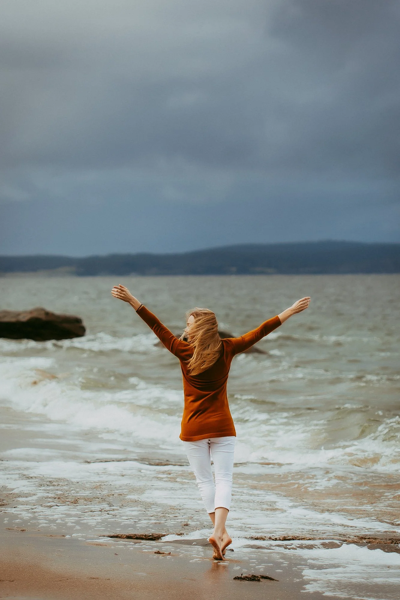 A woman with long hair walking along the beach with her arms raised, facing the ocean under a cloudy sky.