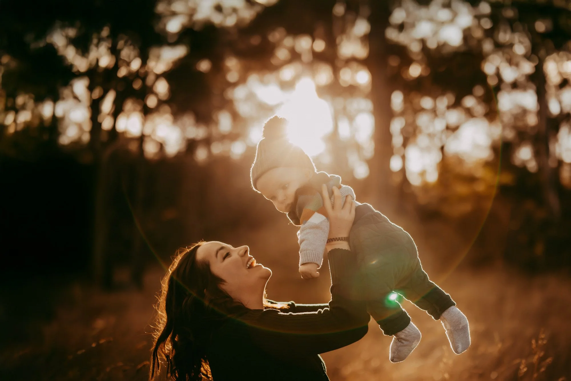 A woman lifting a young child in the air outdoors during sunset, with trees and sunlight in the background. Golden hour family photosession in Greater Hobart area .