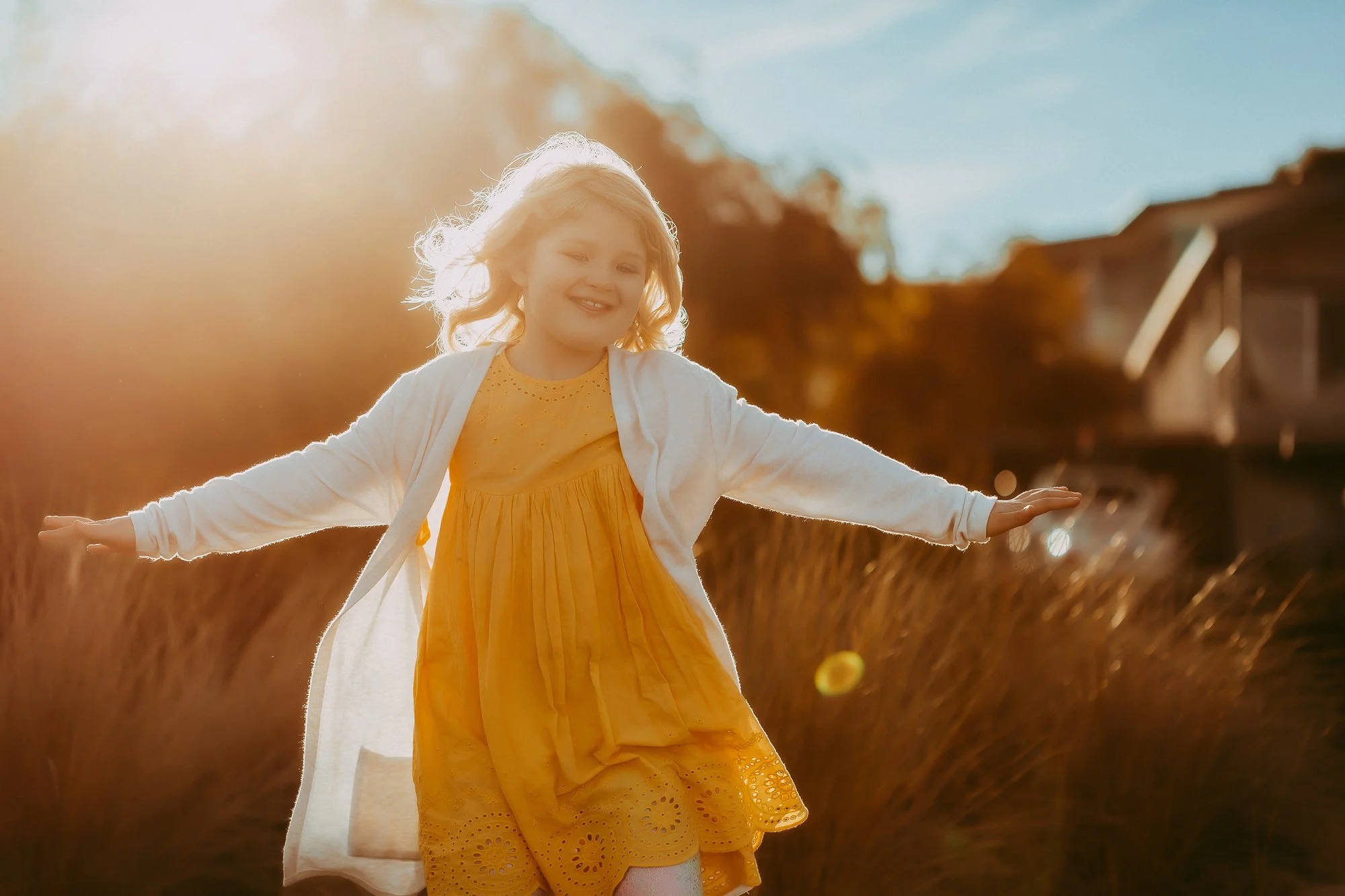 A young girl with blonde hair wearing a yellow dress and white cardigan, smiling and playing in a field during sunset. Golden hour family photosession in Greater Hobart area.