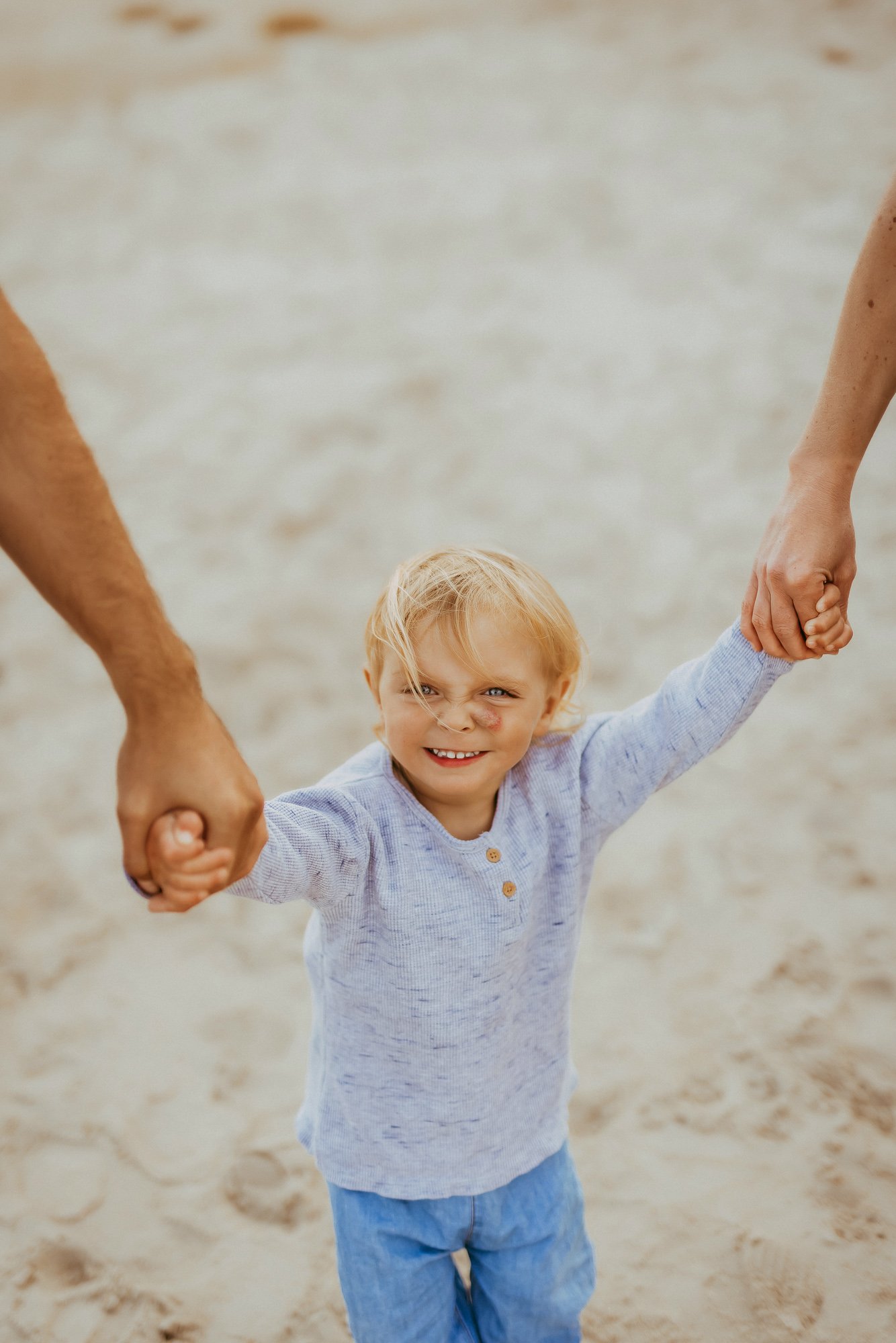 A young boy with blonde hair smiling, holding hands with two adults on a sandy beach.