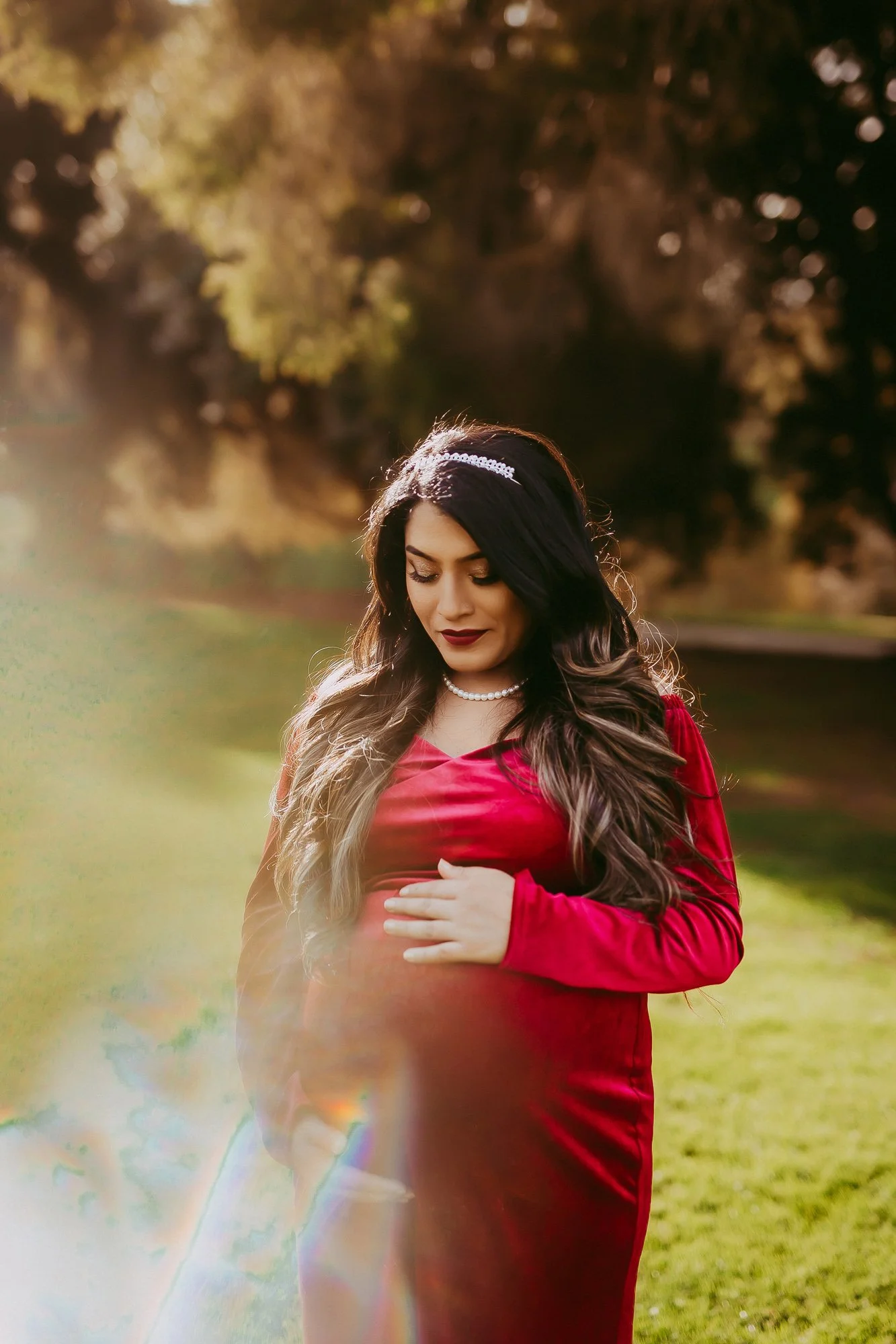 A pregnant woman in a red dress standing outdoors with trees in the background, gently holding her belly and looking down.