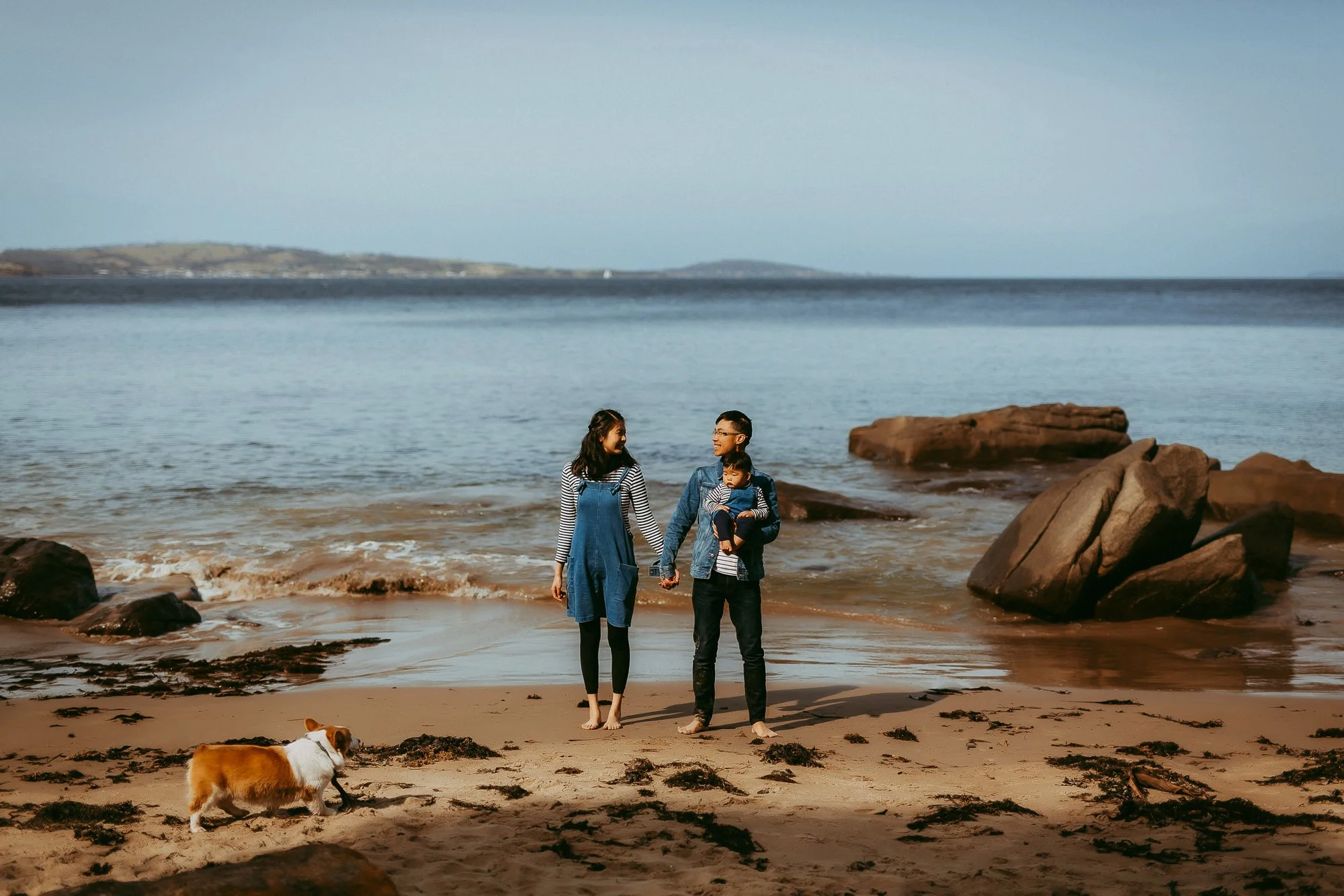 A family of three with a dog walking on a beach during daytime, with rocks and water in the background.