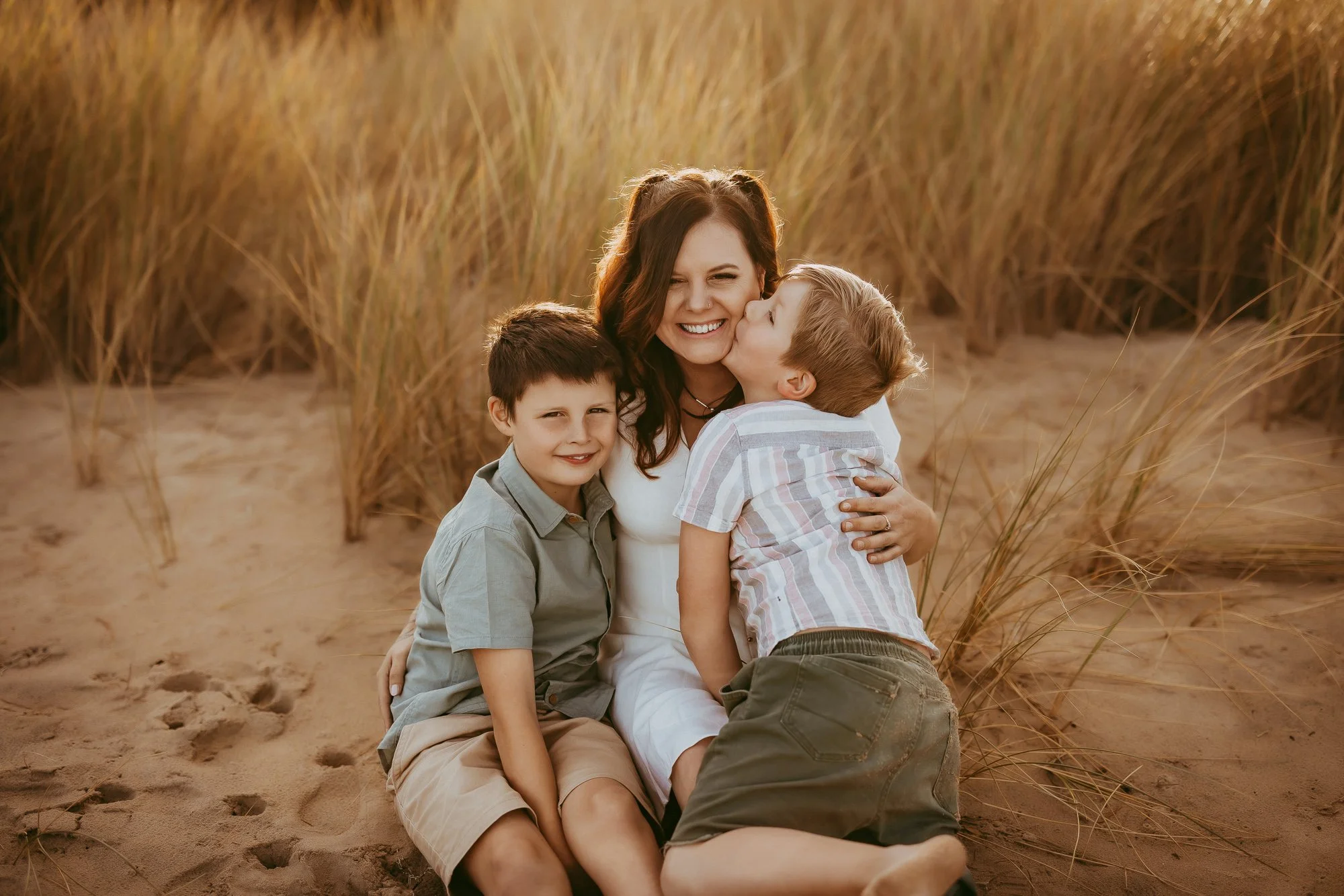 A woman with two young boys sitting on sandy ground among tall grass dunes, one boy giving her a kiss on the cheek, all smiling, during sunset.