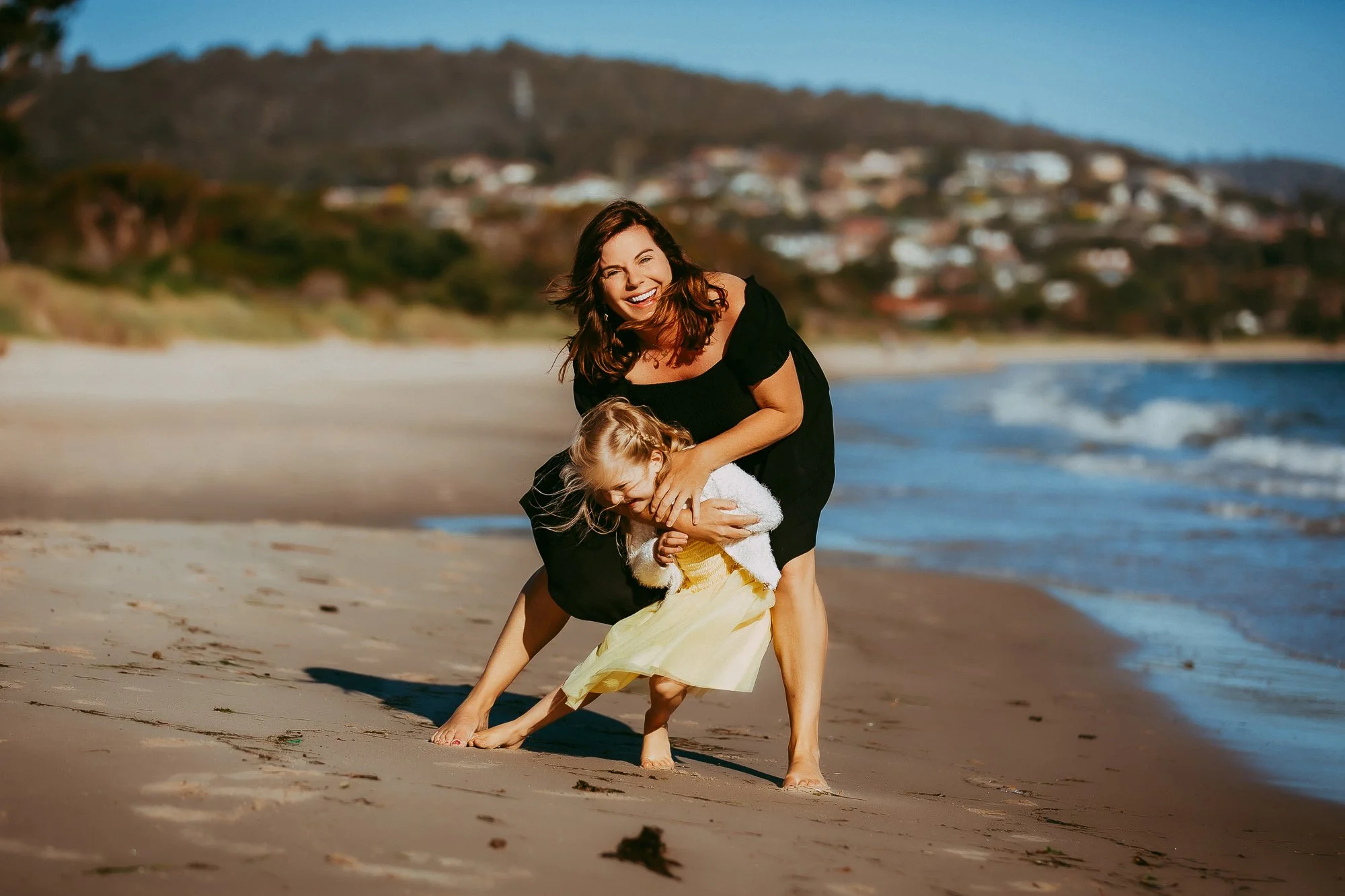 Woman and young girl playing and laughing on the beach with ocean waves and hills in the background. Tasmanian Beach family photography session by Ulla.