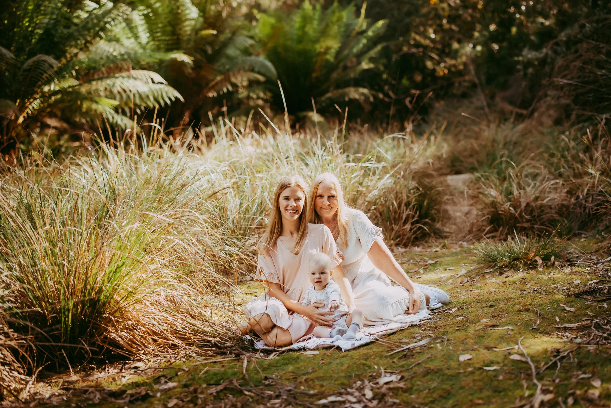 Three generations of women sitting on a blanket in a lush outdoor setting. An elderly woman, a young woman, and a baby girl, smiling at the camera surrounded by tall grasses and greenery.