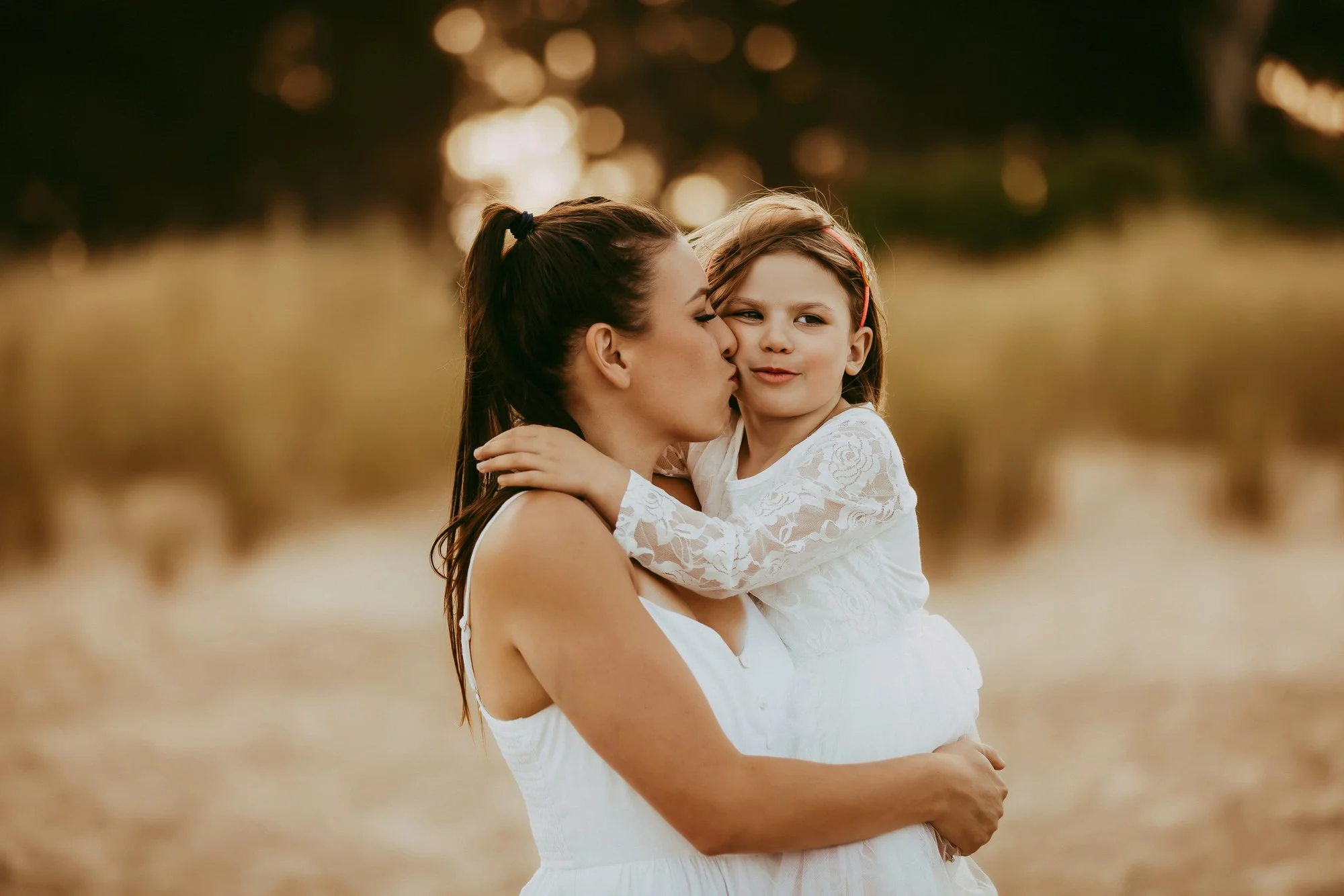 A woman kissing a young girl on the cheek in an outdoor setting during sunset.