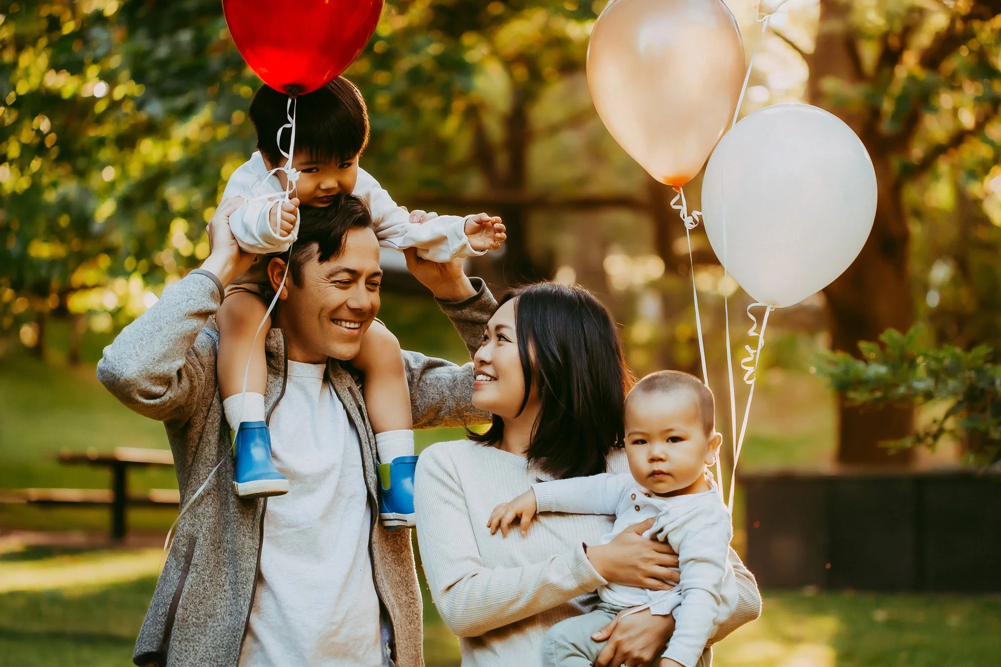 A family of four celebrating outdoors with balloons, a father carrying a child on his shoulders, a mother holding another child, all smiling and enjoying the sunny day. Affordable family photographer at Hobart area.