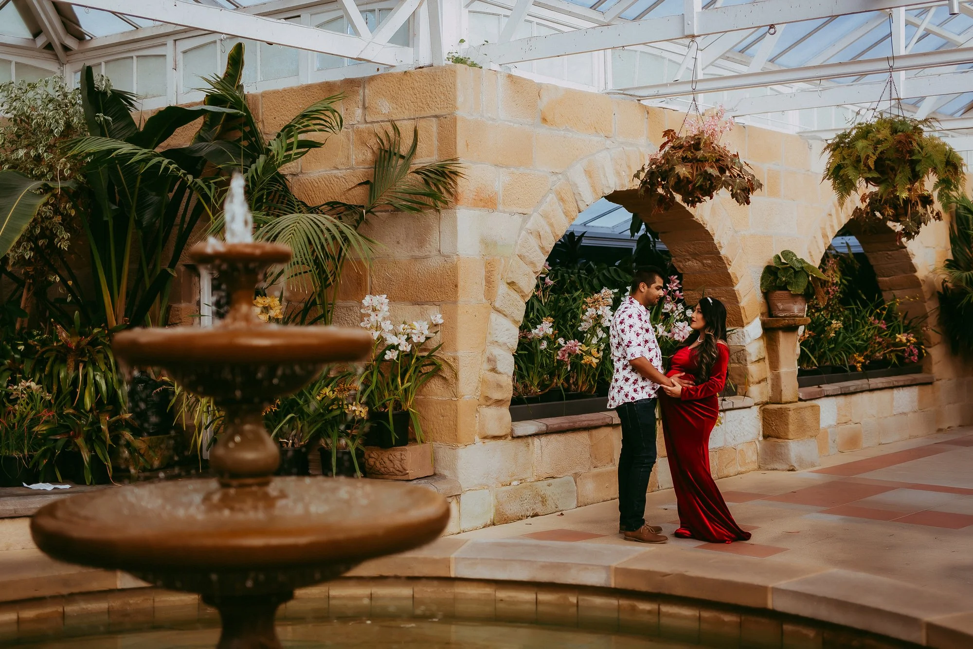 A couple stands holding hands in a lush greenhouse with stone archways, surrounded by green plants and colorful flowers, with a fountain in the foreground.