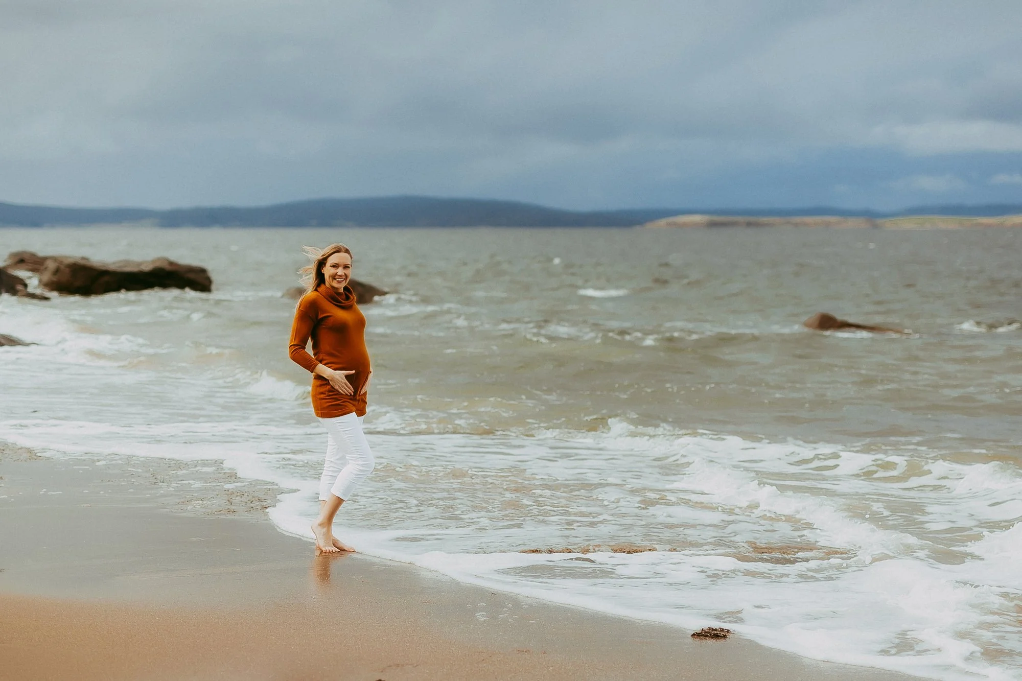 A woman standing barefoot on the beach near the water, smiling and looking at the camera, with short hair and wearing an orange top and white pants, under a cloudy sky.