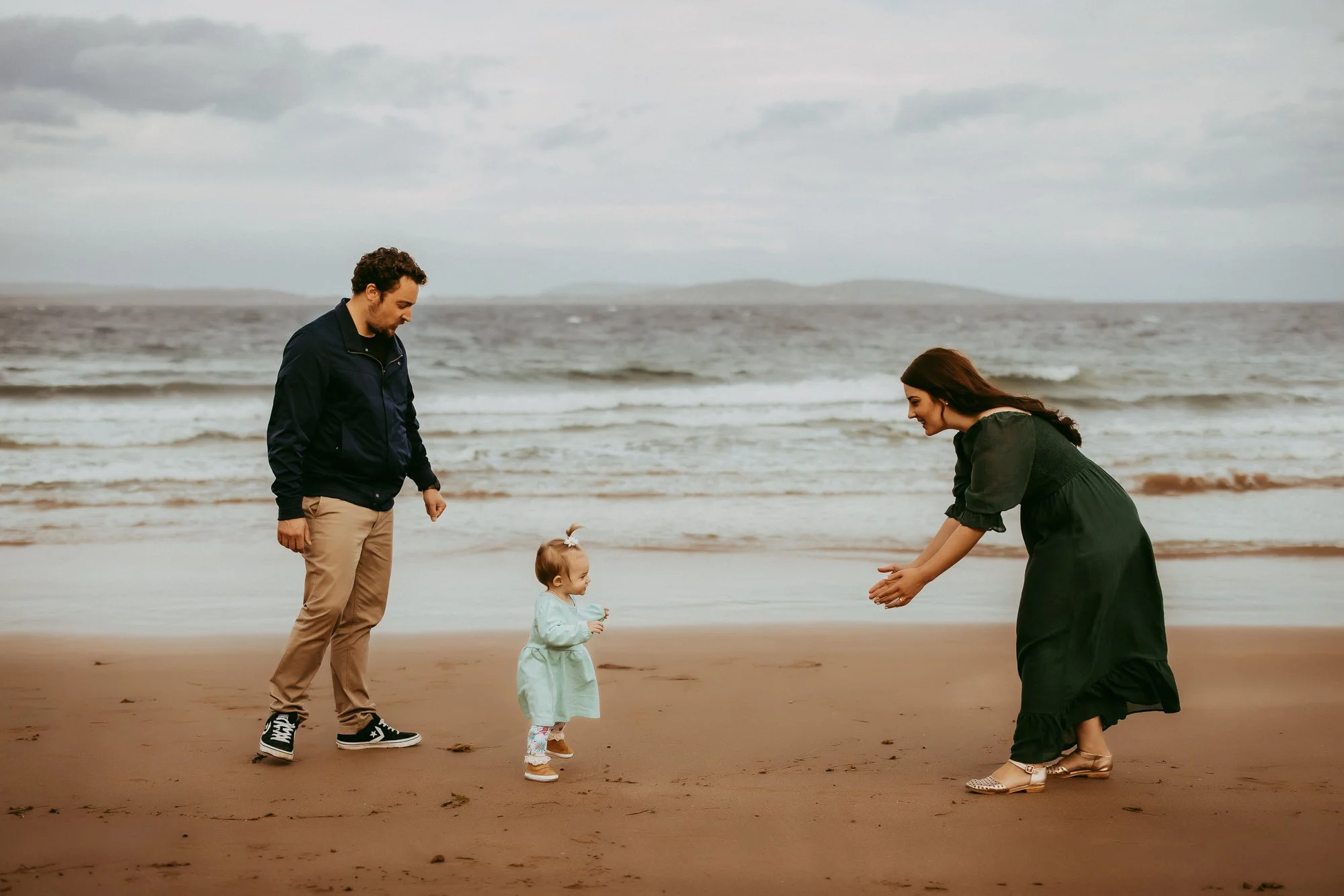A family of three on the beach with the ocean and cloudy sky in the background. The father stands to the left, dressed in beige pants and a navy jacket. The mother is to the right, dressed in a green dress and appearing to blow a kiss or cheer. The t