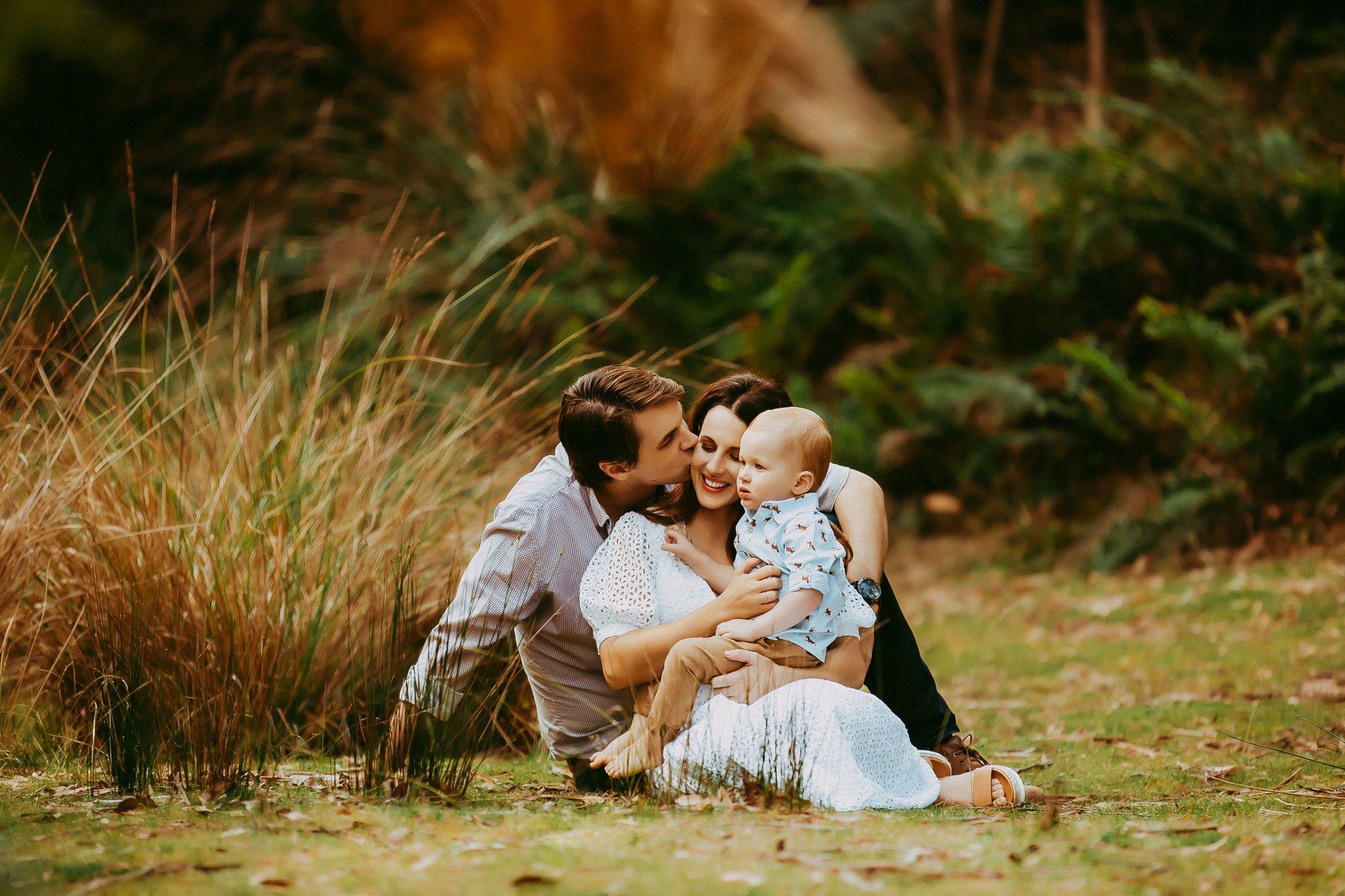 A family of three, a man, a woman, and a young boy, sitting together outdoors in a park-like setting with green foliage and dry grass, sharing a joyful moment, with the man kissing the woman's cheek. Waterworks Reserve family photo session.