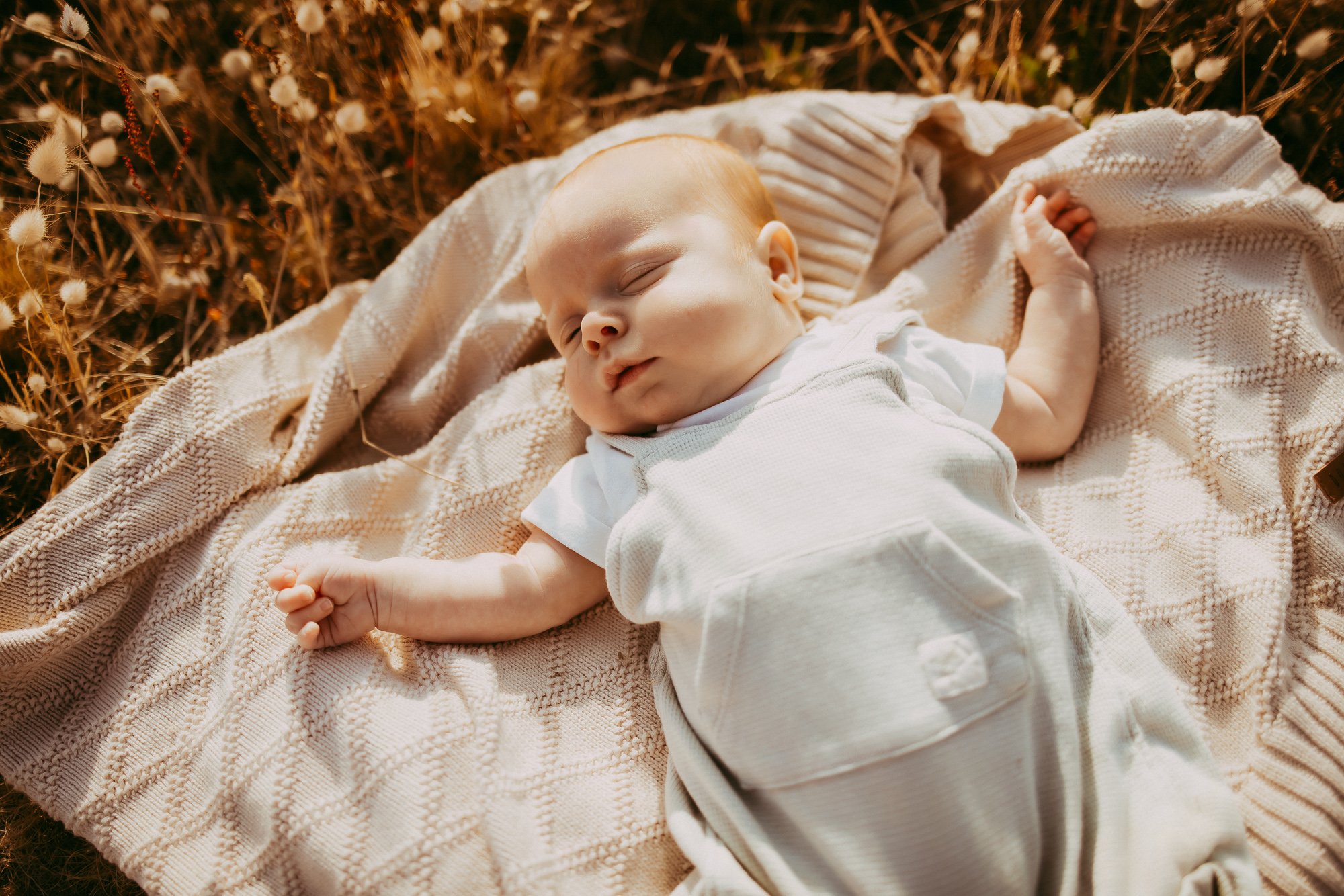 A sleeping baby lying on a beige knitted blanket outdoors in a field of grass and wildflowers Stunning Tasmanian family photo session.