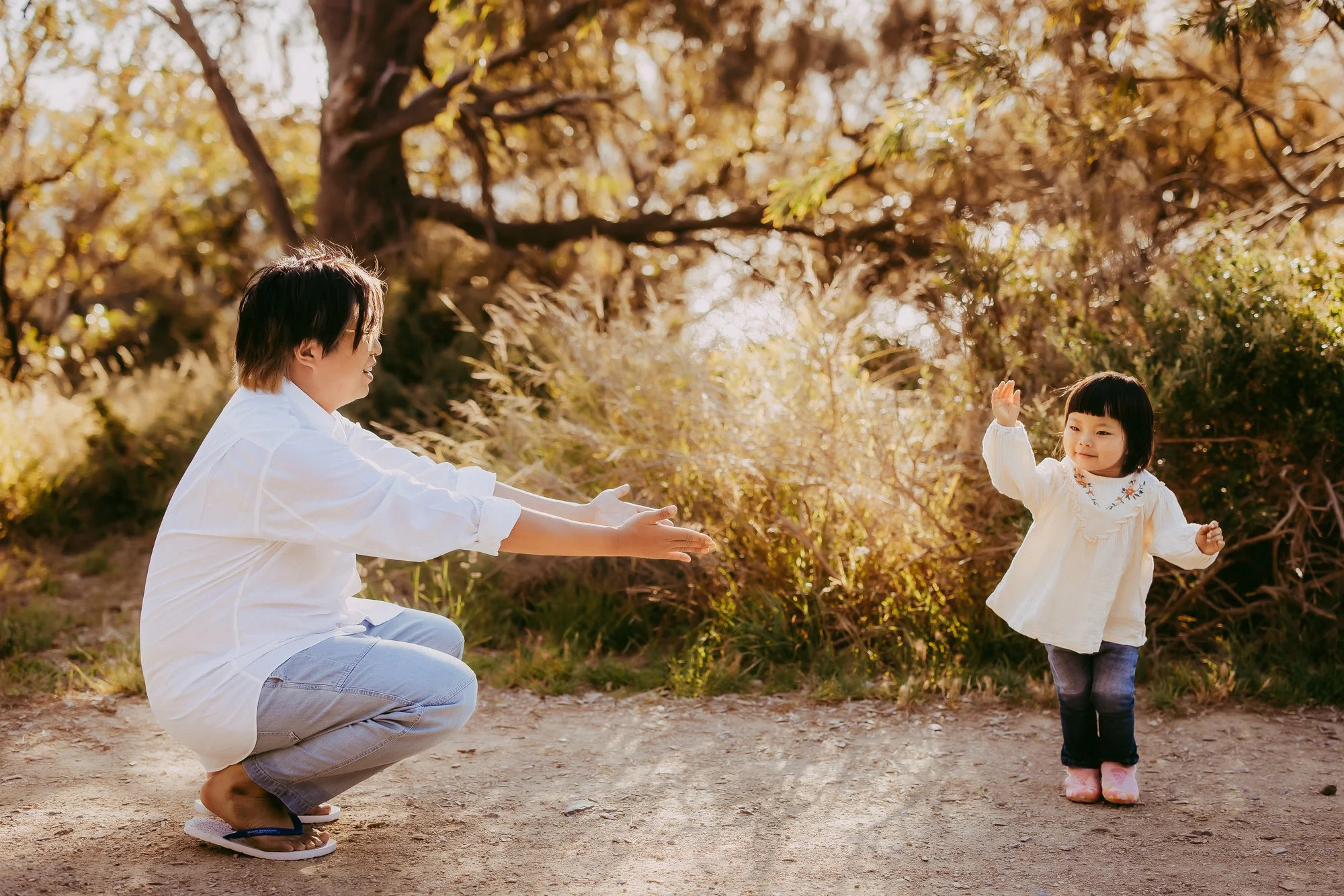An adult crouching with arms extended toward a young girl standing on a dirt path, waving, with a background of trees and shrubs in warm sunlight. Golden hour family photography session in Hobart Tasmania.