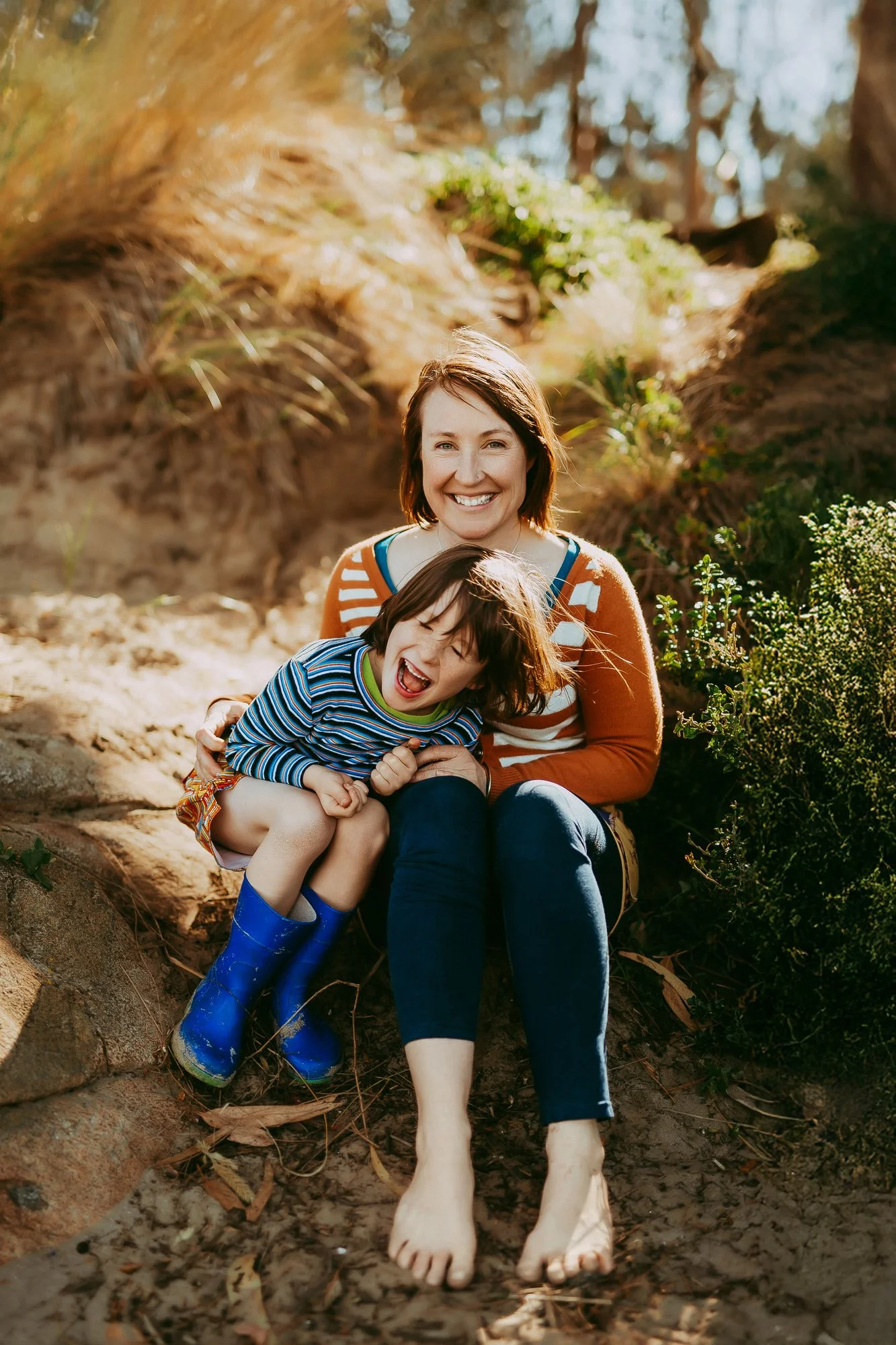 A woman and a young girl sitting on a sandy outdoor area, smiling and laughing together. Greater Hobart family photography session with relaxed approach.