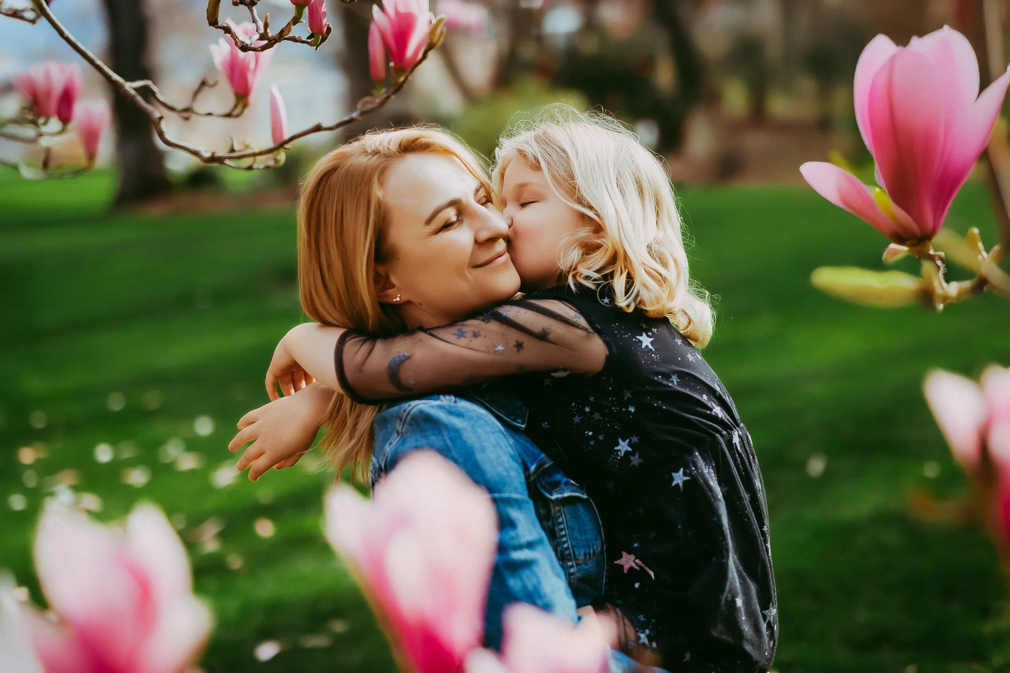 A woman and a young girl embrace and share a kiss outdoors among blooming pink magnolia flowers.