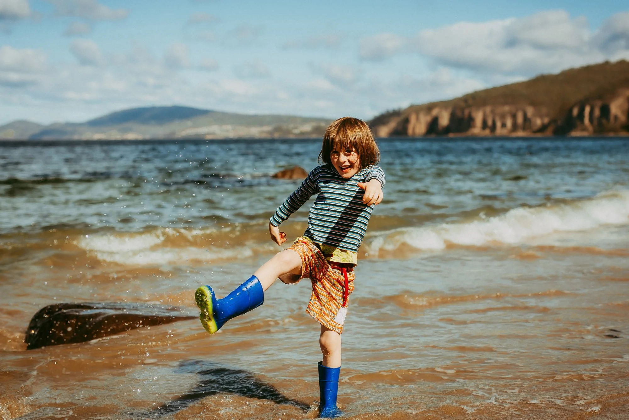 Child playing in the water on a beach, wearing rain boots and smiling. Affordable family photographer at Hobart area.