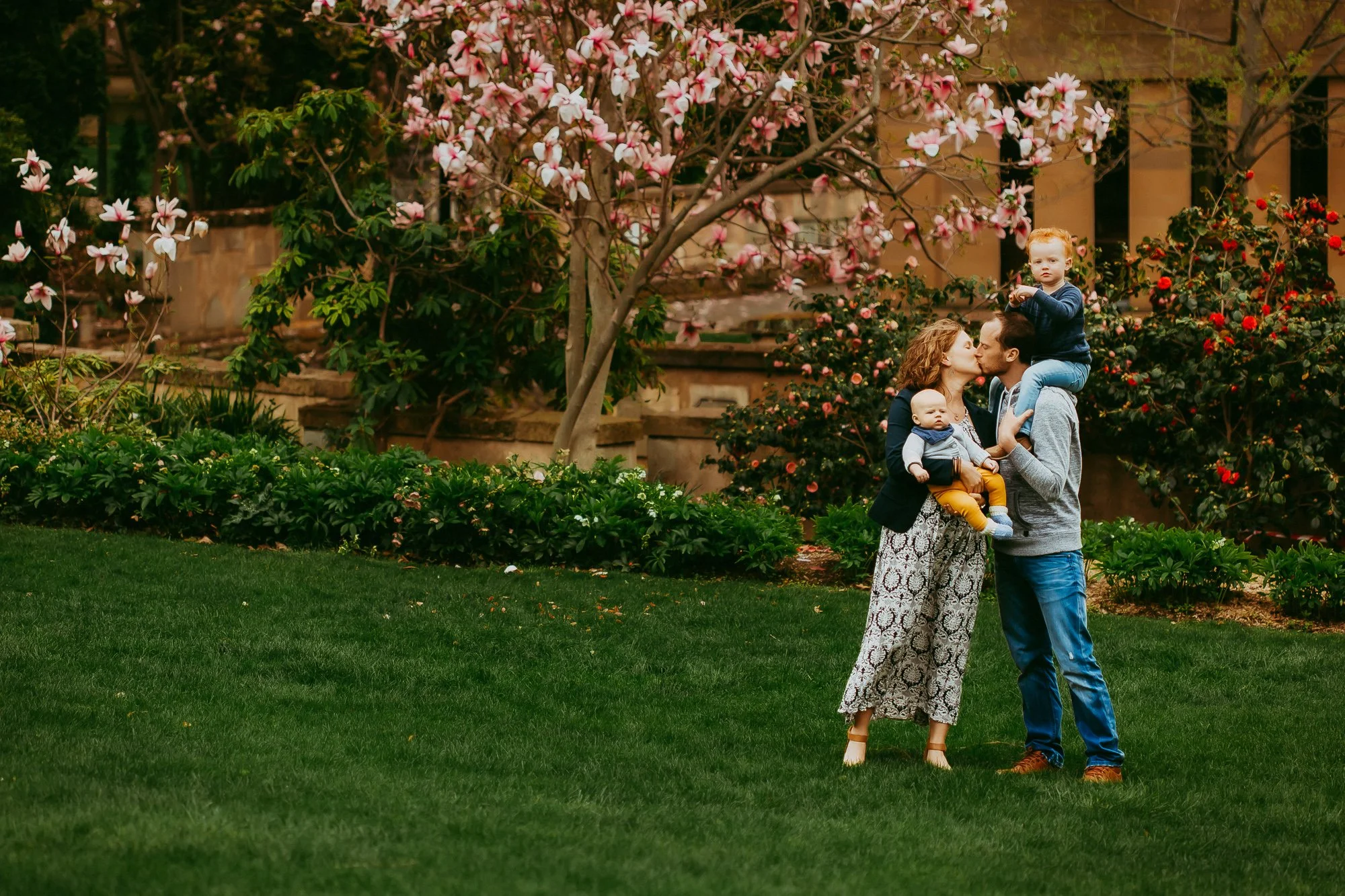 A family of four with two young children, a woman, and a man, standing on a lush green lawn in a garden with blooming pink and red flowers. The woman and man are kissing, and one child is sitting on the man's shoulders while the other child is held b