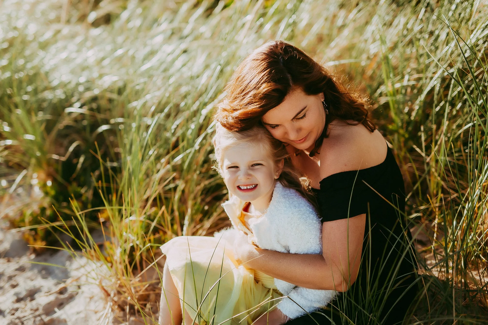 A woman and a young girl sitting on a grassy area, smiling and enjoying the sunlight. Greater Hobart family photographer.