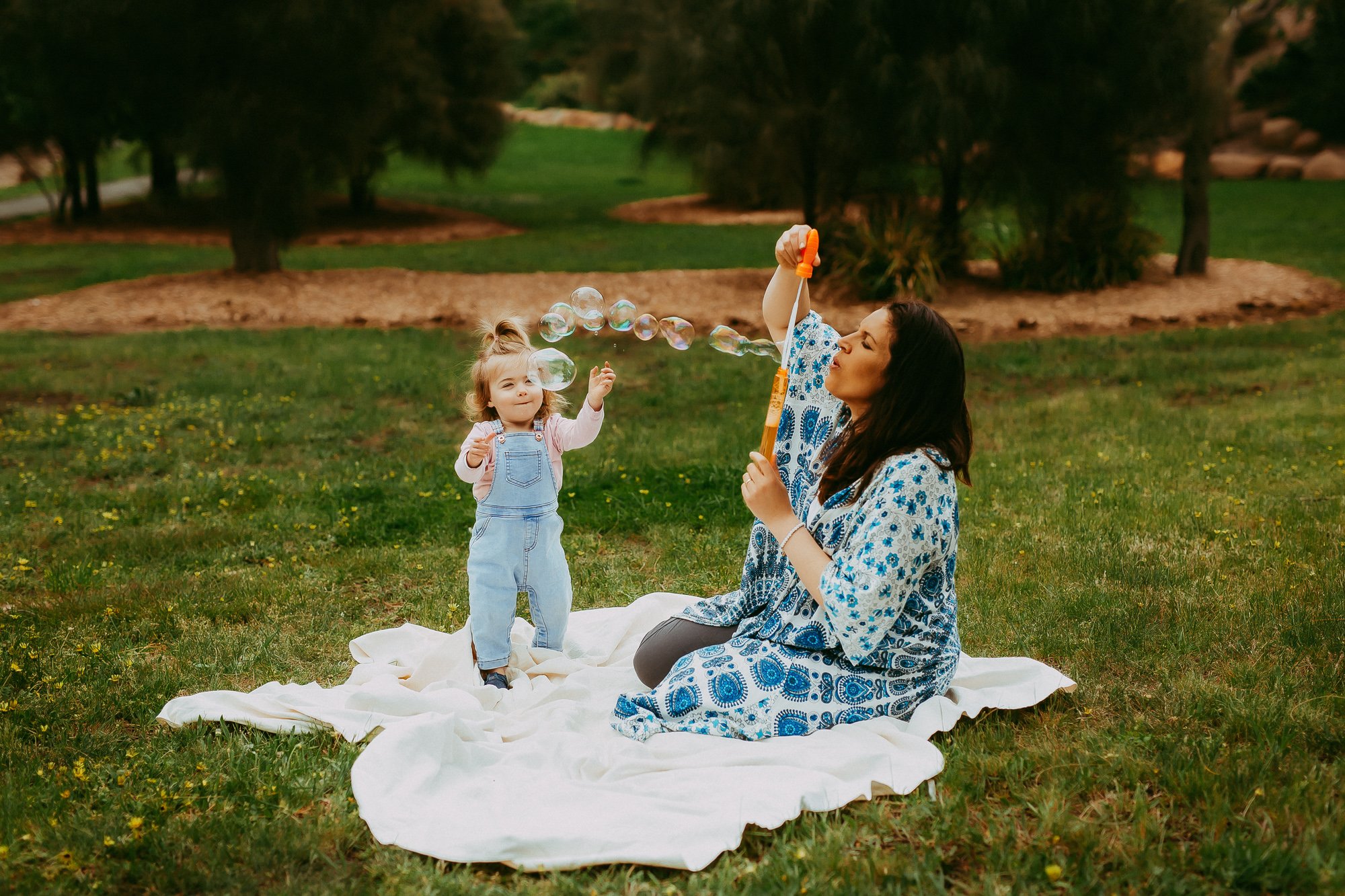 Woman sitting on a white blanket blowing bubbles while a young girl in denim overalls watches, outdoors in a grassy park with trees in the background.