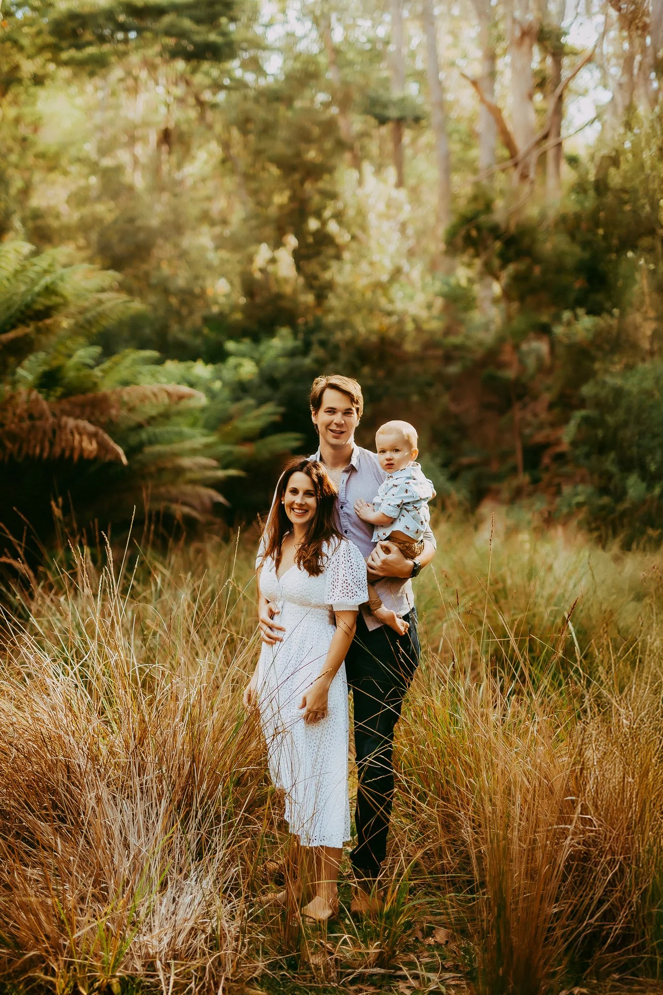A family of three standing in a field of tall grass with dense trees in the background, during golden hour.