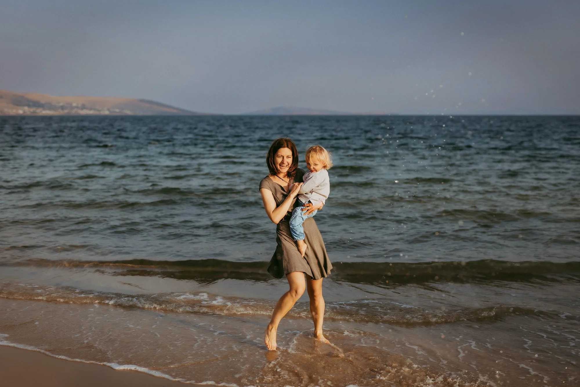A woman and a young child playing and smiling at the beach, standing in shallow water with waves, with hills in the background.