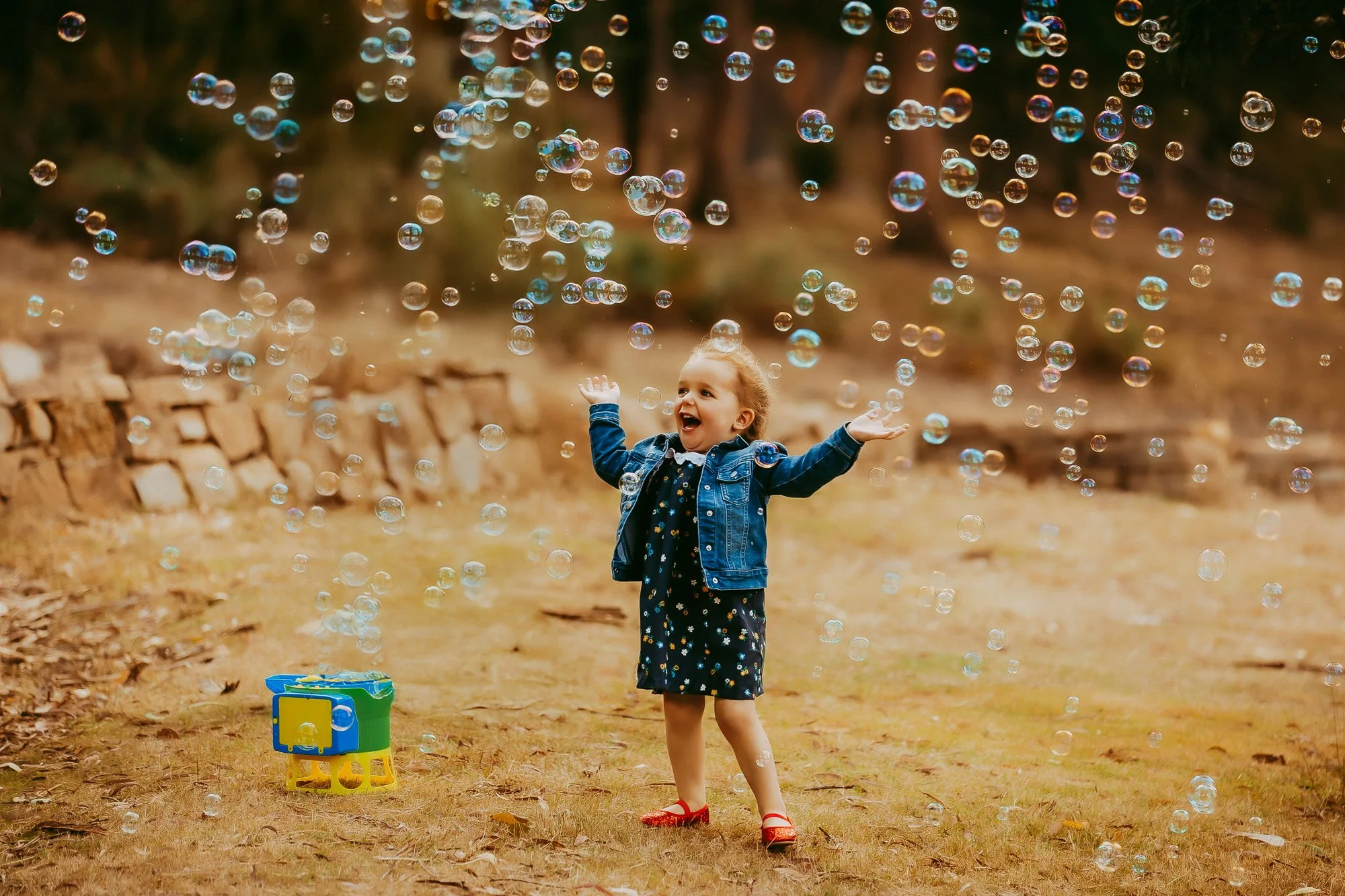 A young girl in a black dress, red shoes, and a denim jacket playing with bubbles outdoors on a grassy area with a stone wall and trees in the background.