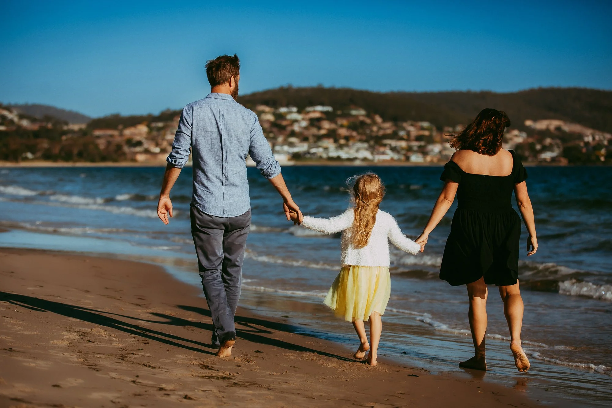 A family of three walking hand in hand on a sandy beach near the water. Golden hour family photo session in Greater Hobart area.