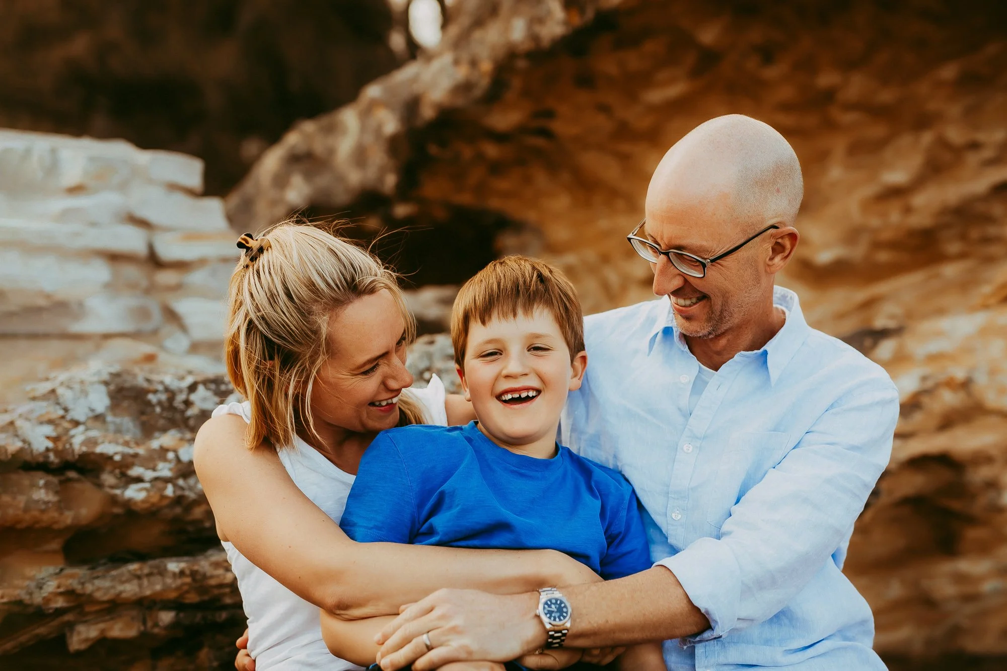 A happy family of three, a woman, a man with glasses, and a young boy, smiling and embracing outdoors with rocks and a natural setting in the background.