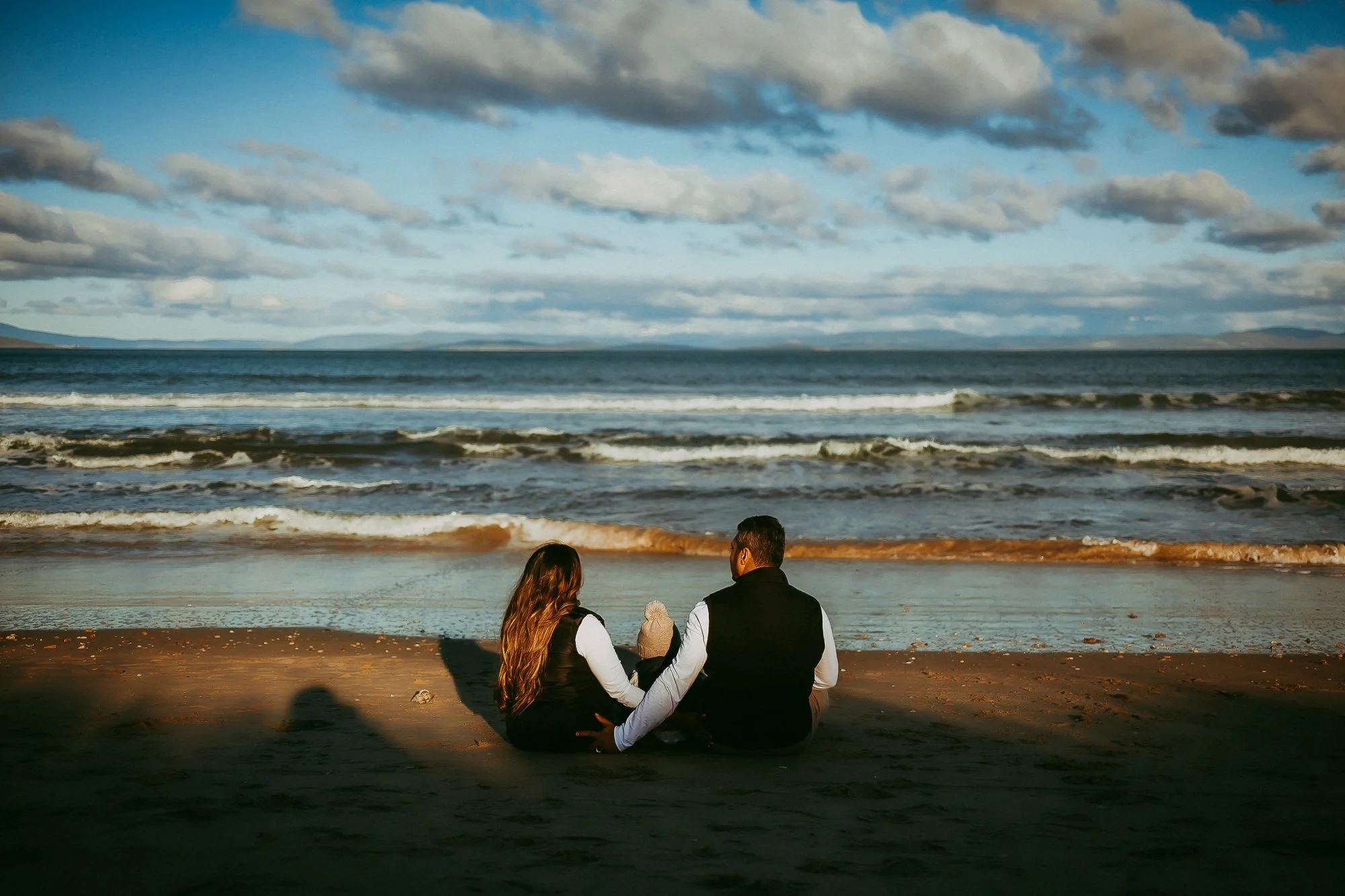 A couple sitting on the beach facing the ocean, with overcast skies and waves in the background. Tasmanian Beach family photosession.