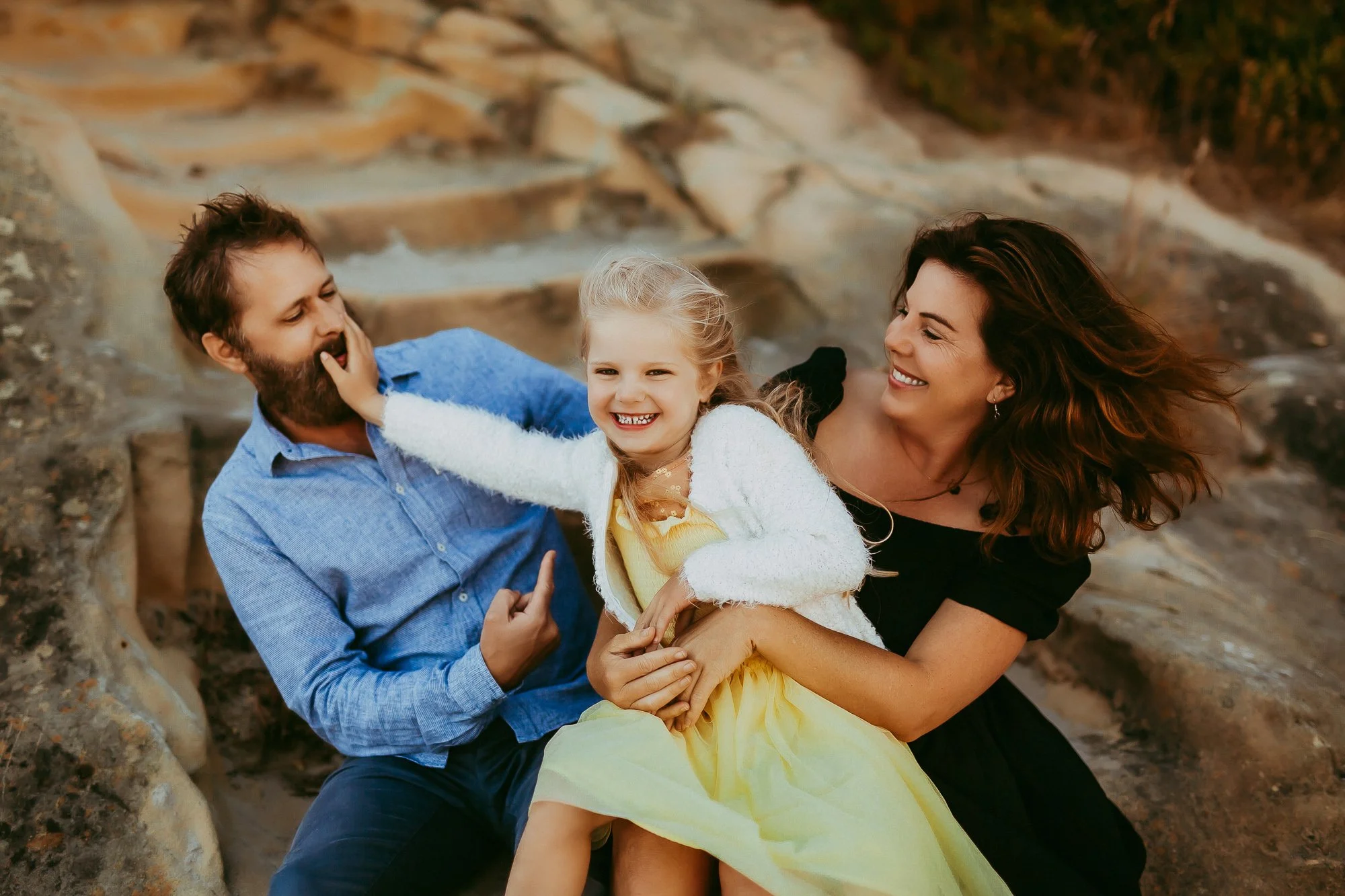 Happy family of three, a man, woman, and young girl, playing and smiling outdoors on rocky terrain. Tasmanian Beach family photosession.