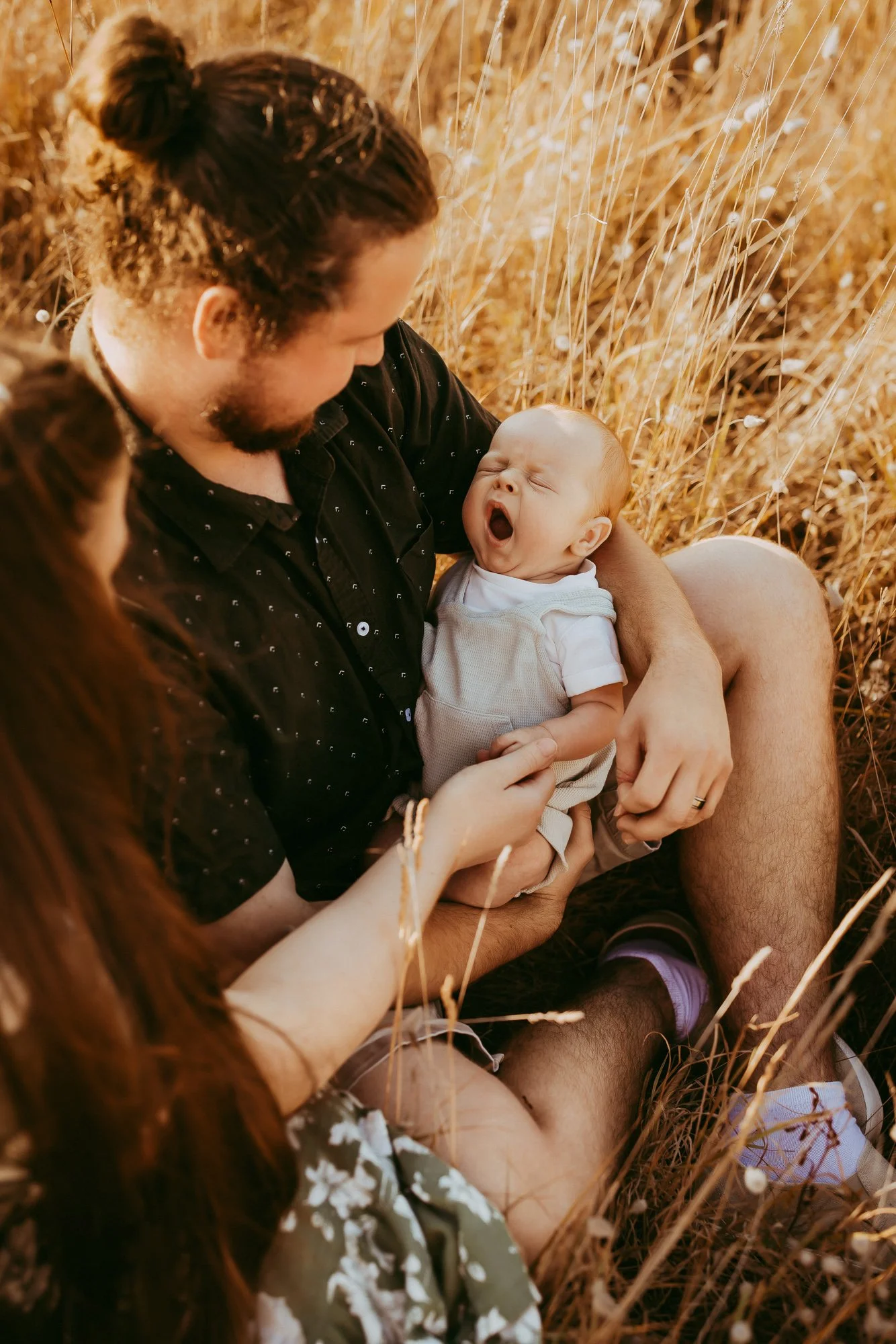 A man holding a yawning baby in a field of tall golden grass, with a woman reaching out towards the baby. Family photosession at Tasmanian Beach.