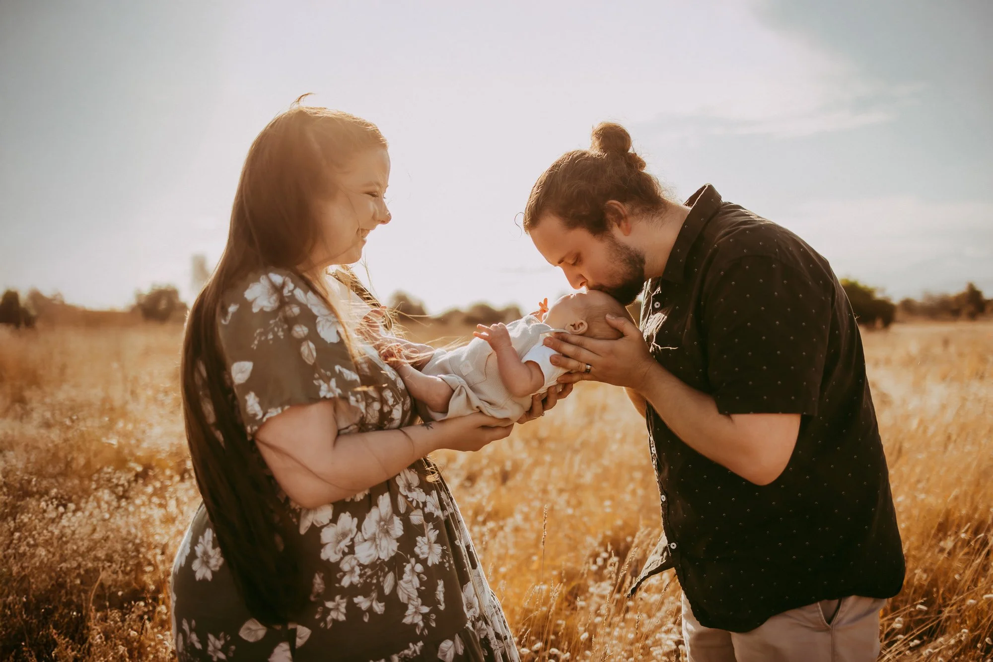 A couple holding their baby in a field during sunset, with the mother smiling and the father kissing the baby's forehead. South Arm Tasmania family photosession at golden hour.