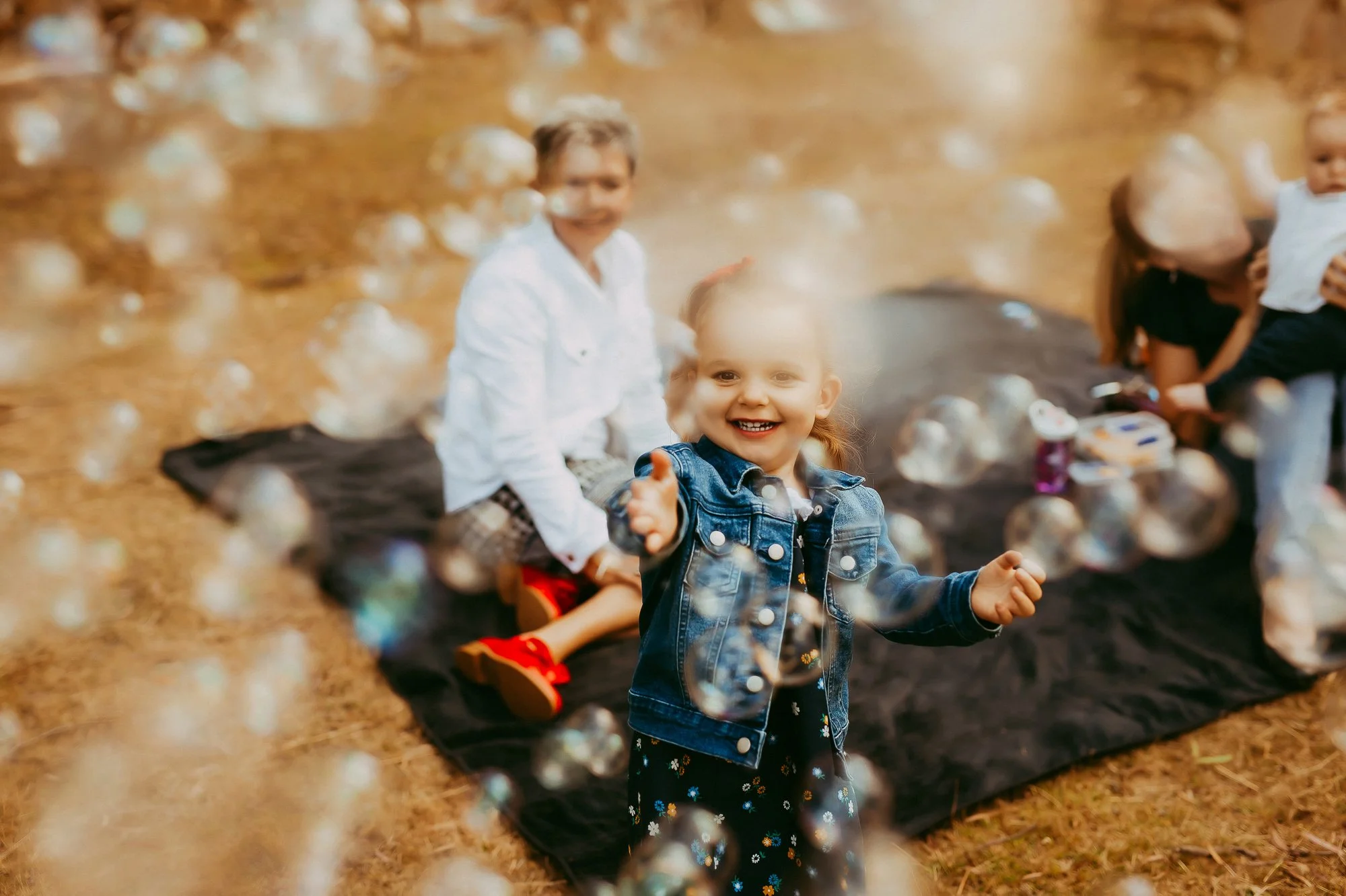 Smiling young girl in denim jacket reaching for bubbles during outdoor gathering with children on black blanket.