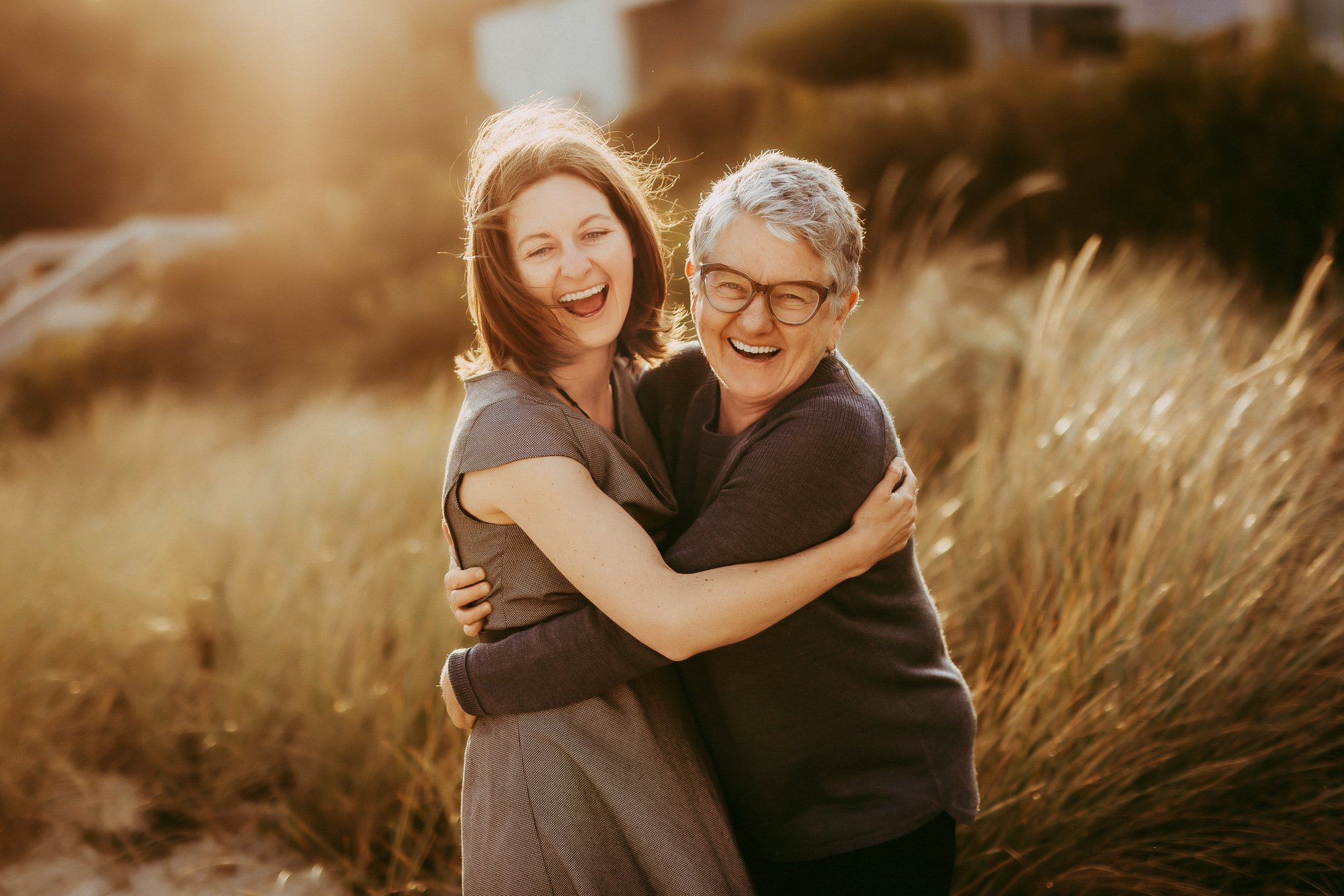 Two women, one with gray hair and glasses and one with red hair, sharing a hug and smiling happily outdoors during sunset with tall grass in the background.