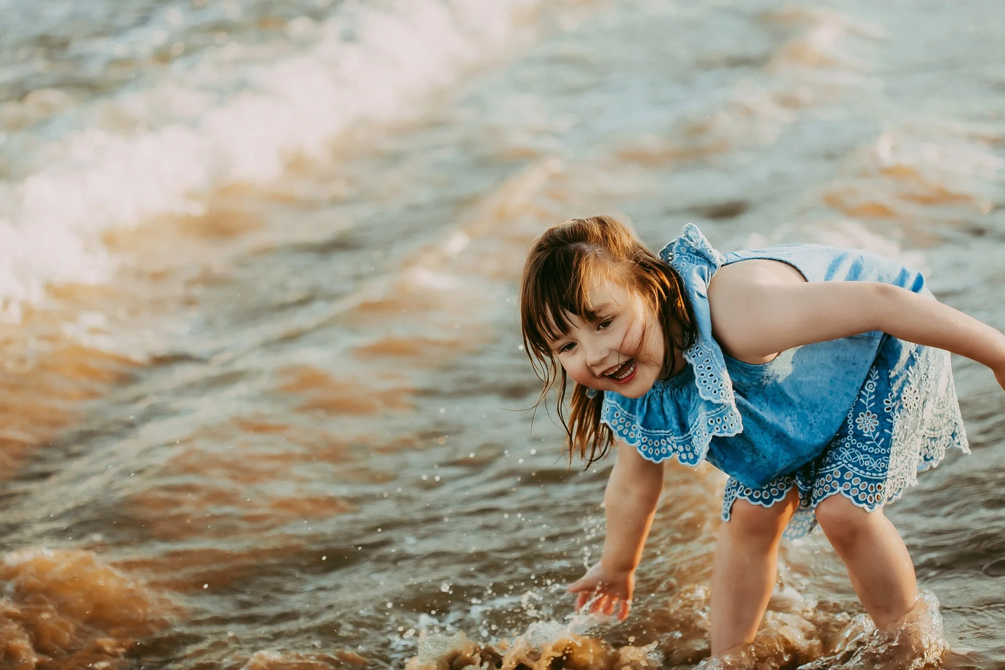 A young girl with brown hair in a blue dress playing in the ocean water at the beach, smiling and reaching into the water. Seven Mile Beach Tasmania. Affordable family photographer Ulla.