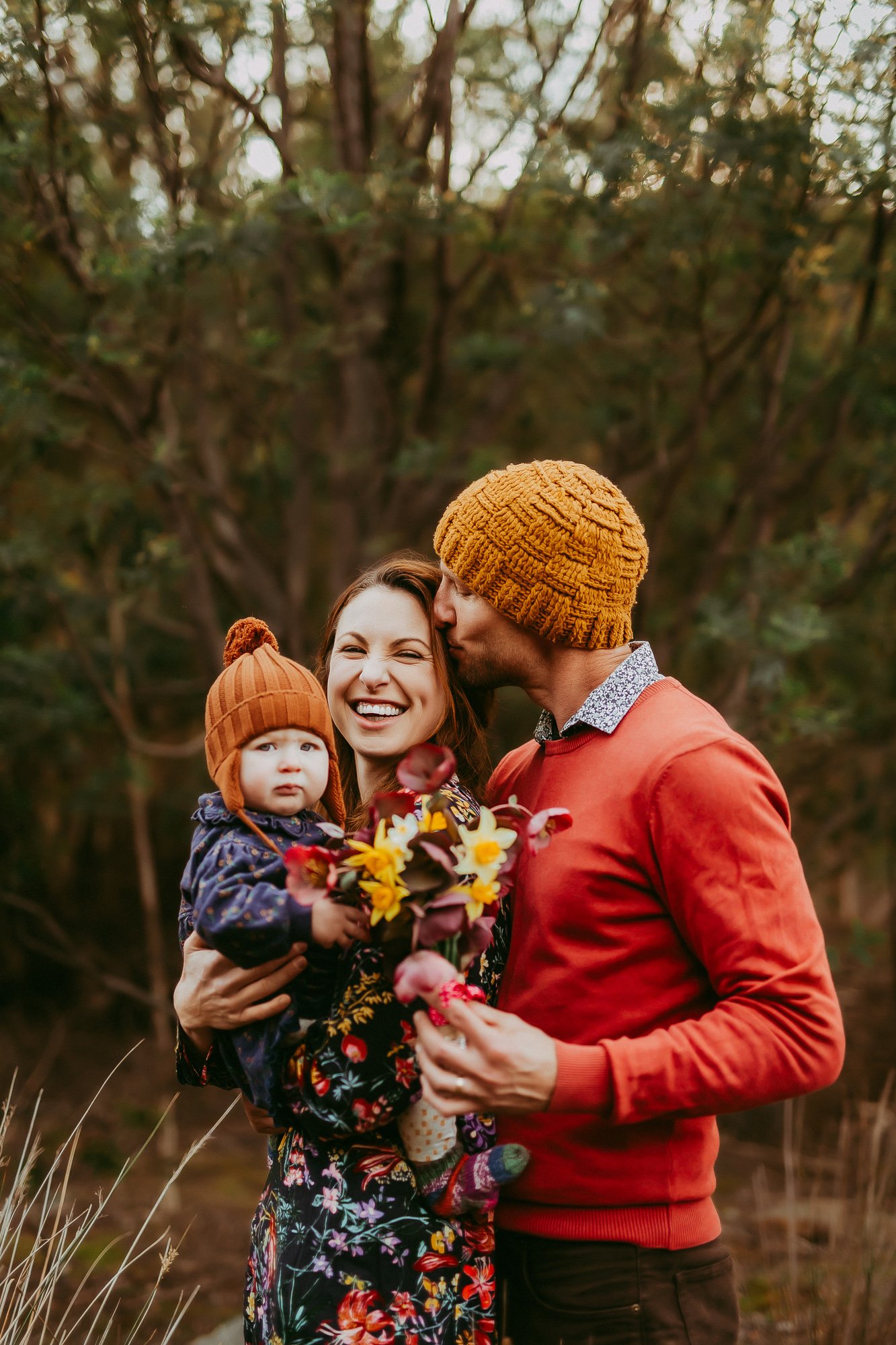Family of three outdoors, woman holding a child, man kissing woman's forehead, weather is cool, autumn background
