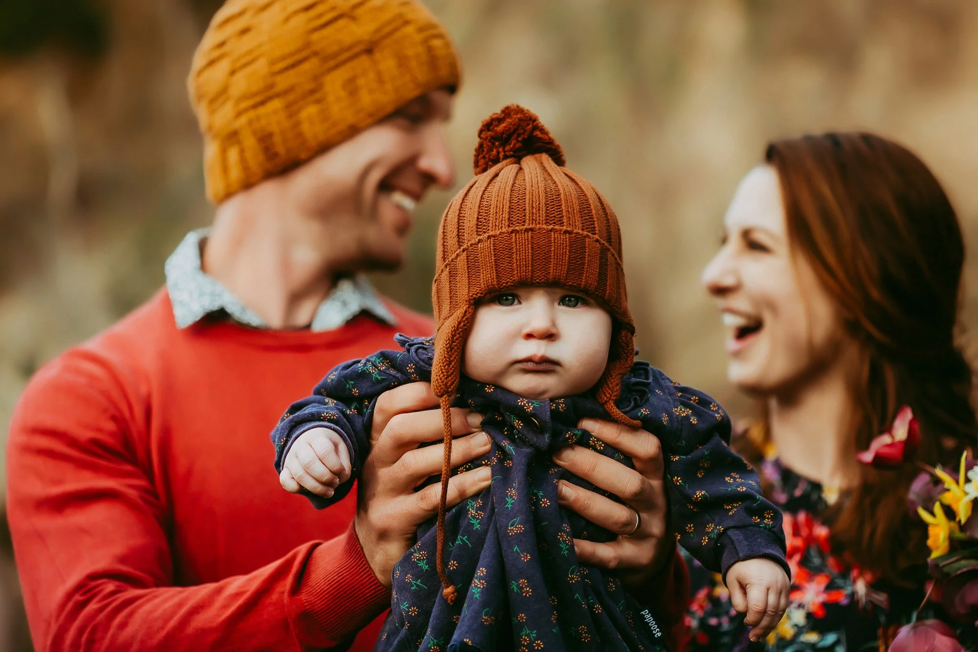 A man and woman holding a baby outdoors during fall. The man wears an orange sweater and a yellow knit hat, and the woman wears a dark floral top. Budget friendly family photosession in Hobart area.