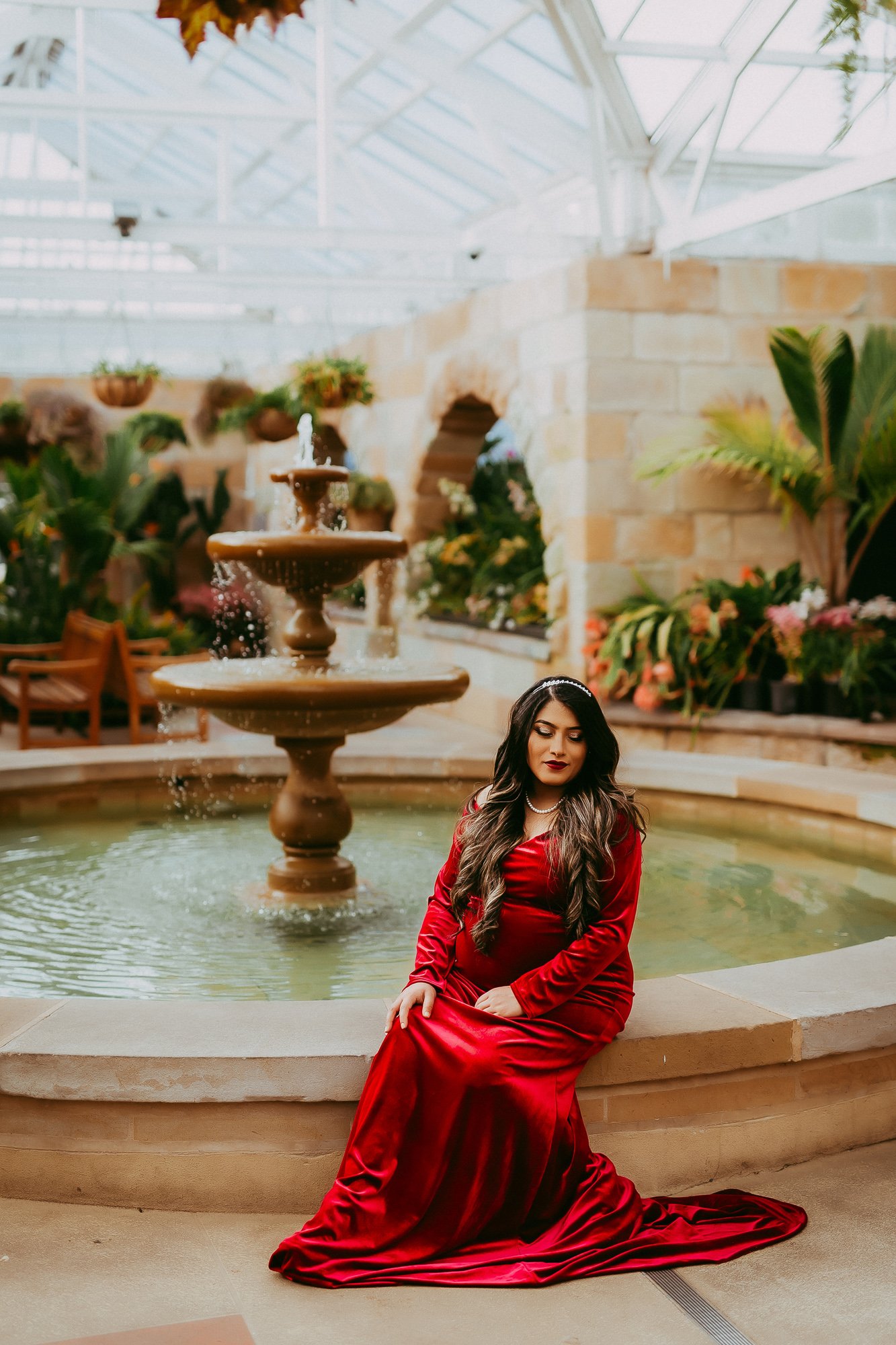 A woman in a red gown sitting by a fountain inside a greenhouse or conservatory filled with plants and flowers.
