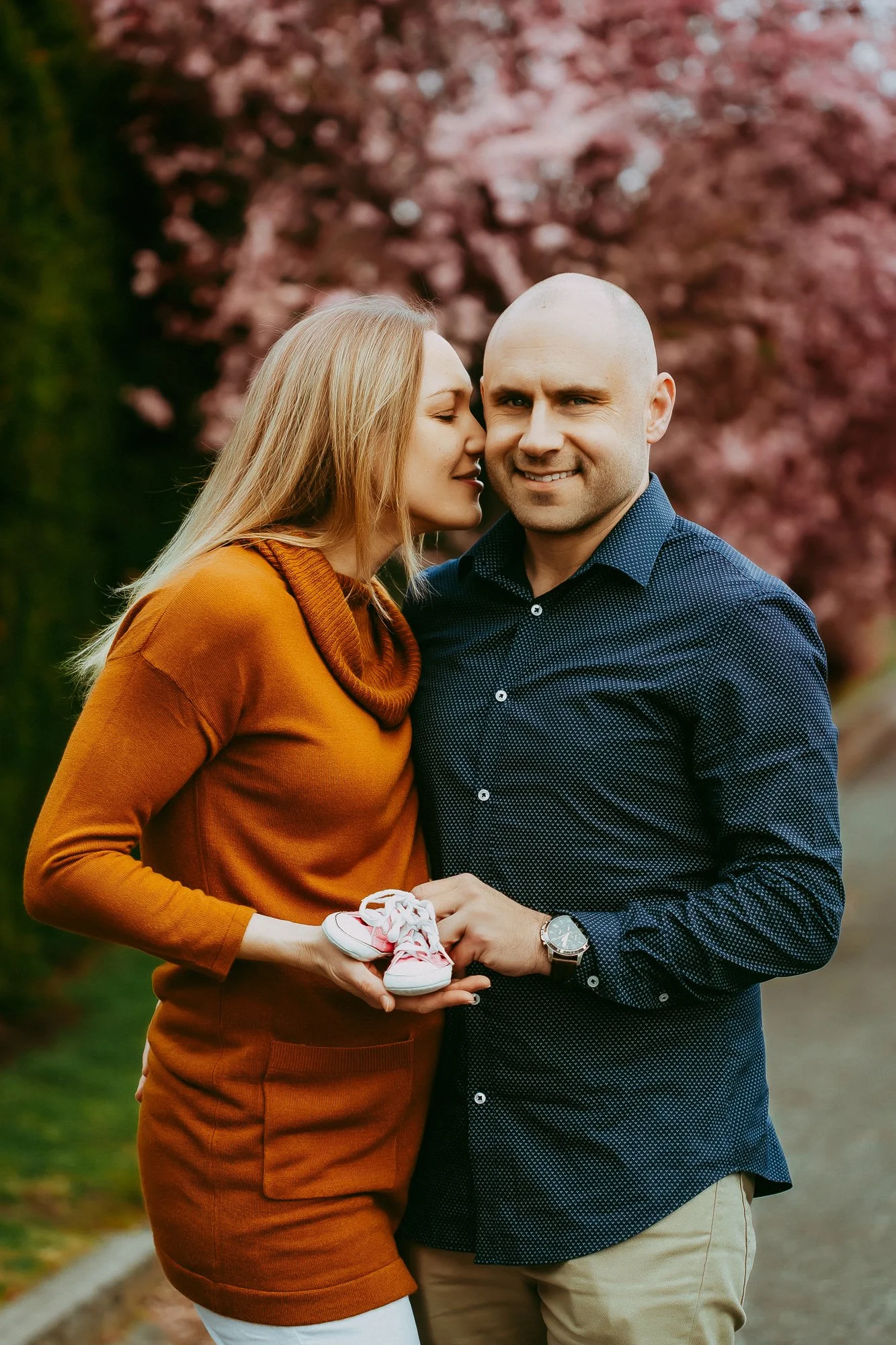 A couple standing outdoors near pink cherry blossom trees, with the woman whispering into the man's ear. The woman is holding a pair of pink sneakers. They are smiling and embracing each other.