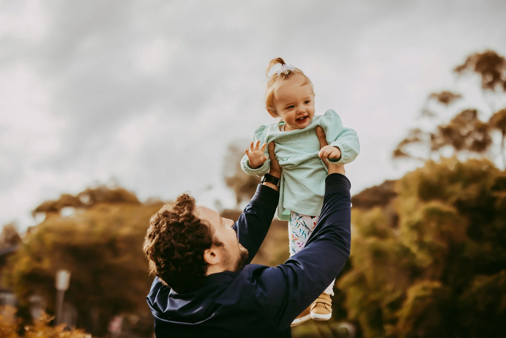 A man lifting a smiling toddler girl in the air outdoors during daytime, with trees and cloudy sky in the background.