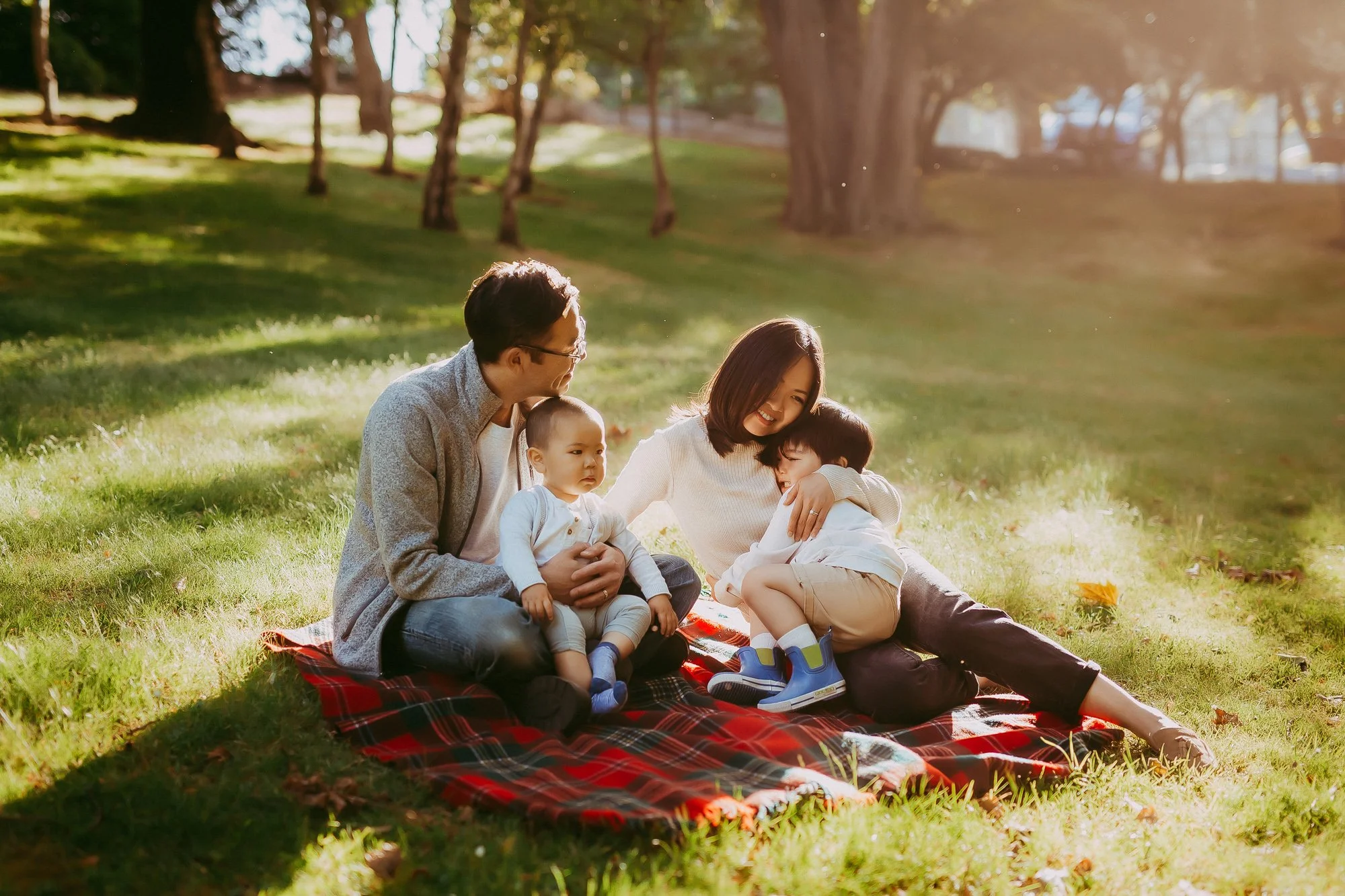A family of four sitting on a red plaid blanket outdoors in a park, with tall trees in the background, enjoying a sunny day. Cascade Brewery family photoshoot.