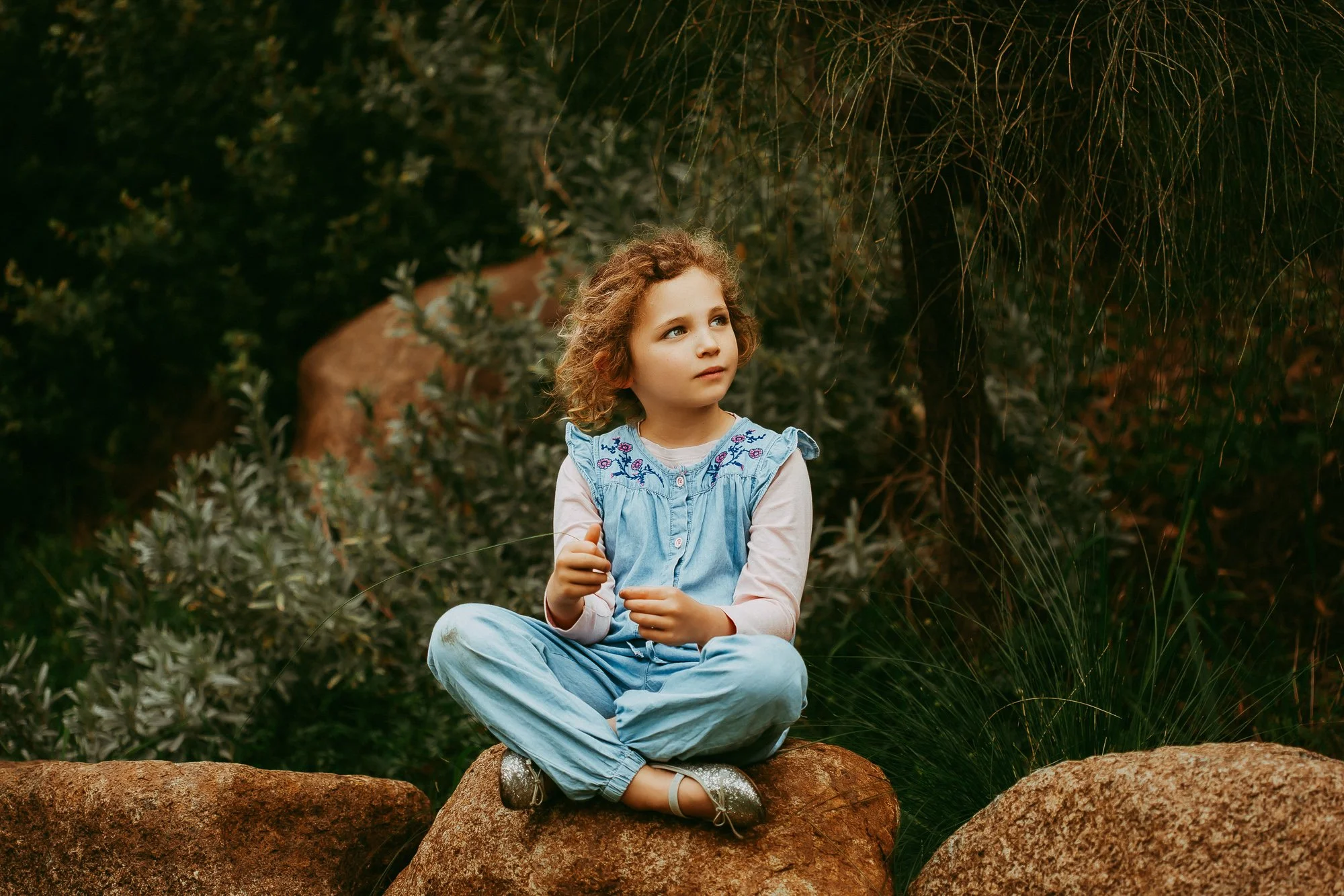 A young girl with curly hair sitting cross-legged on a rock outdoors, surrounded by greenery and rocks, looking thoughtful.