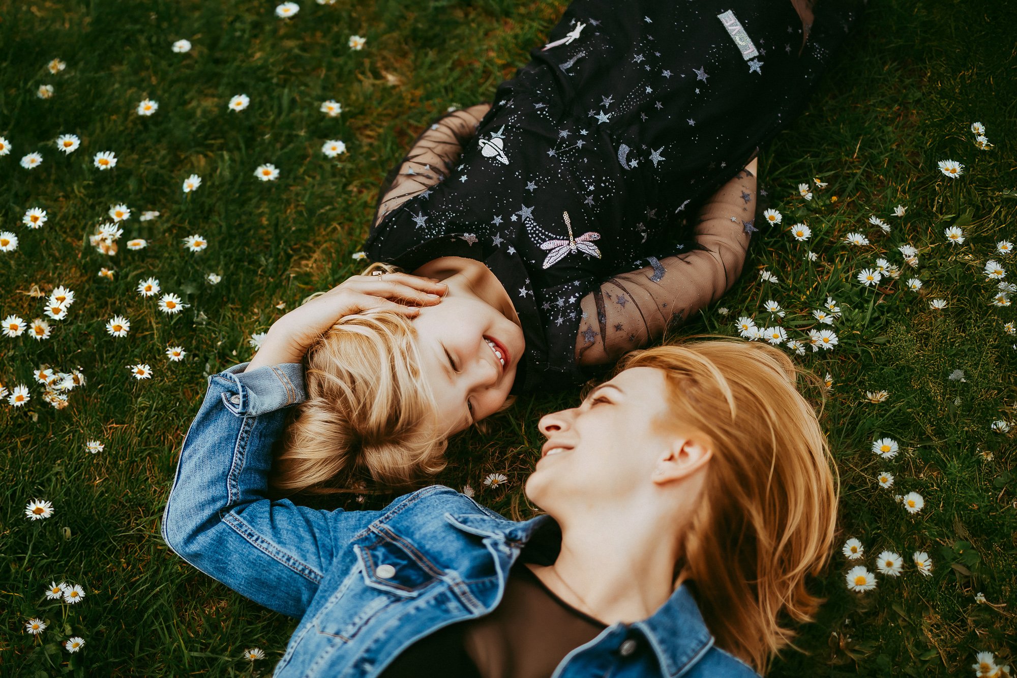 Two young women with blonde and red hair lying on grass with daisies, smiling at each other. Budget friendly relaxed and chill Hobart family photography.